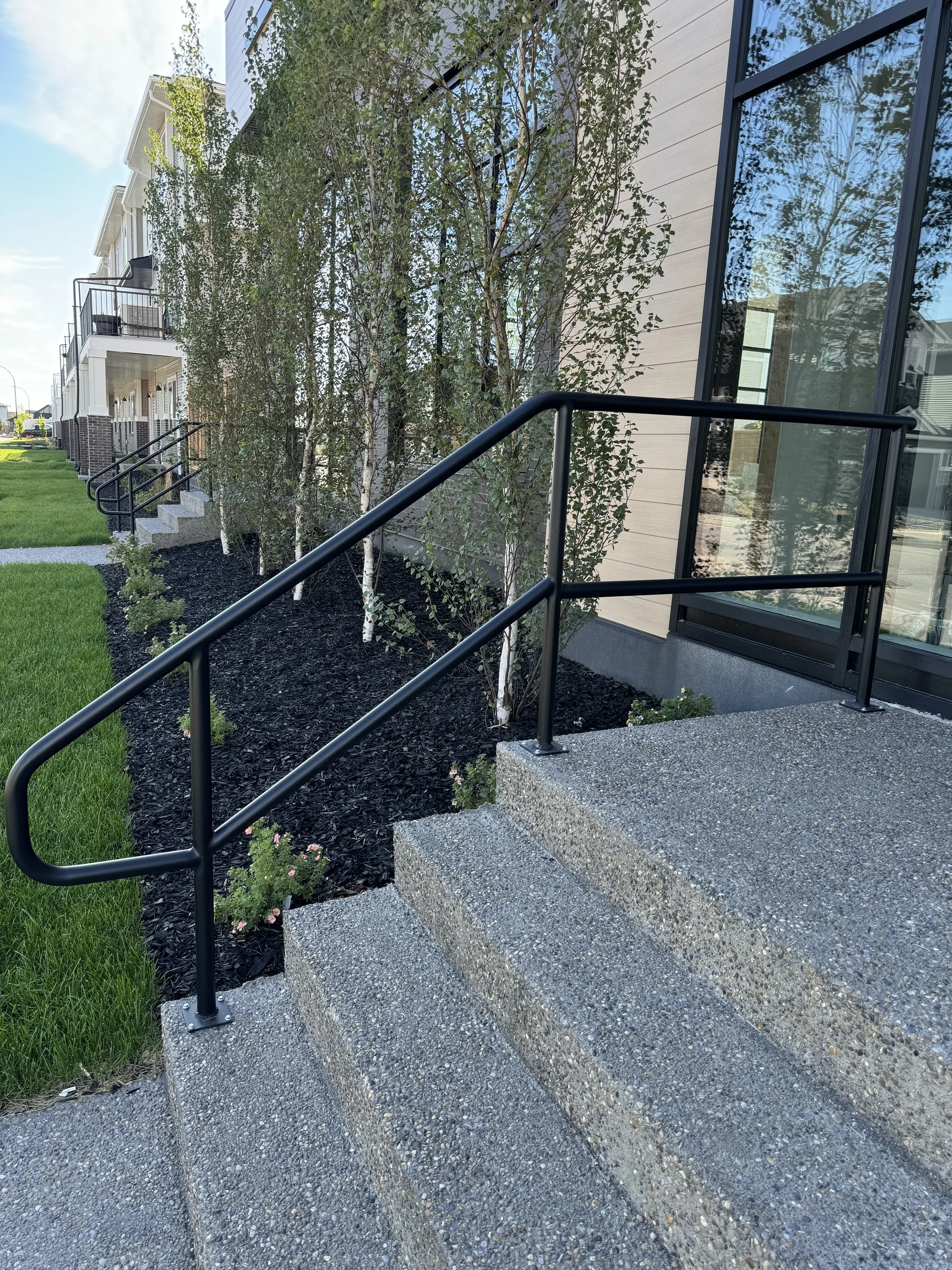 Concrete steps with black metal handrails outside a modern building, with trees and grass nearby.