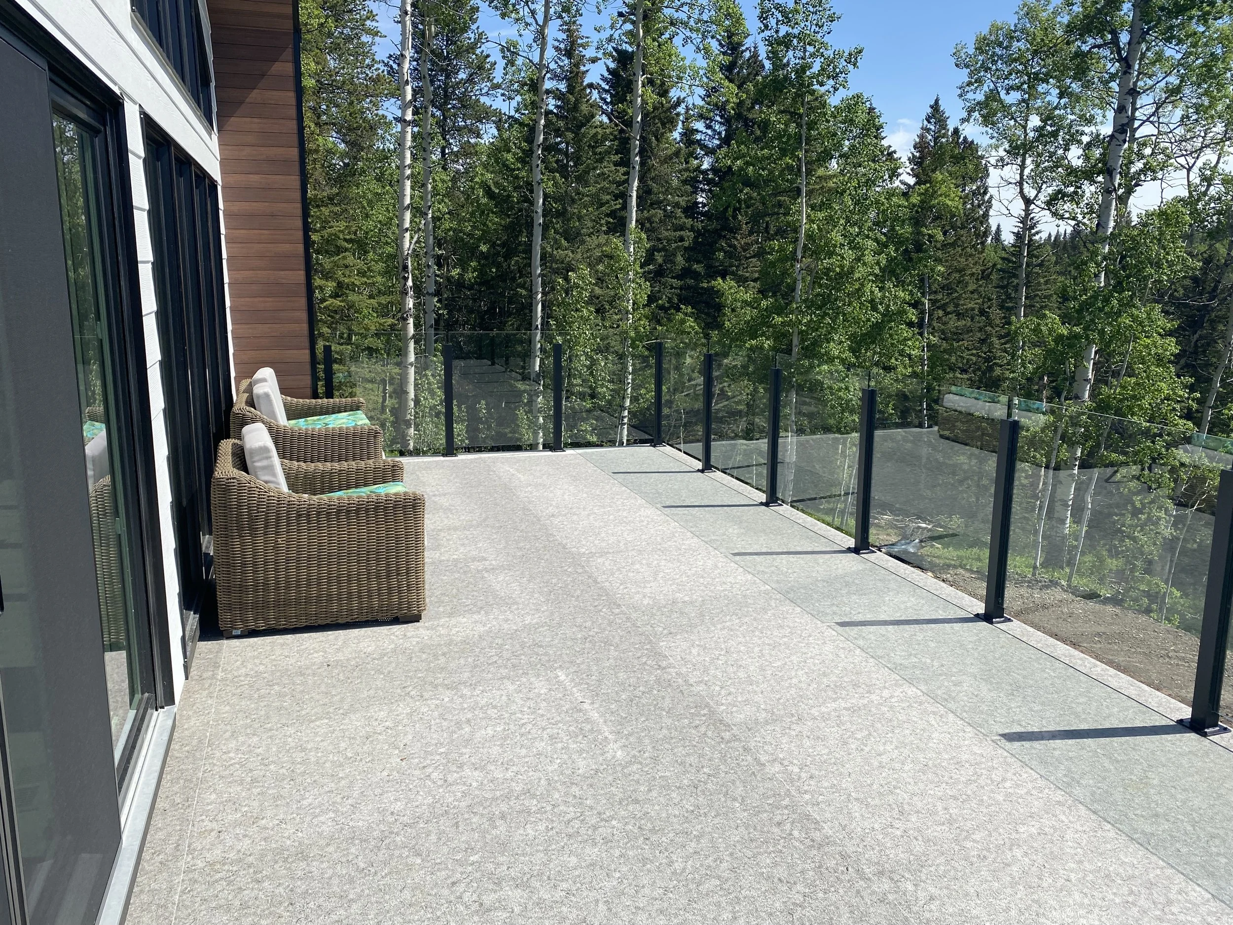 A spacious balcony or terrace area with wicker chairs and cushions, surrounded by a glass railing, overlooking a forest with tall green trees under a clear blue sky.