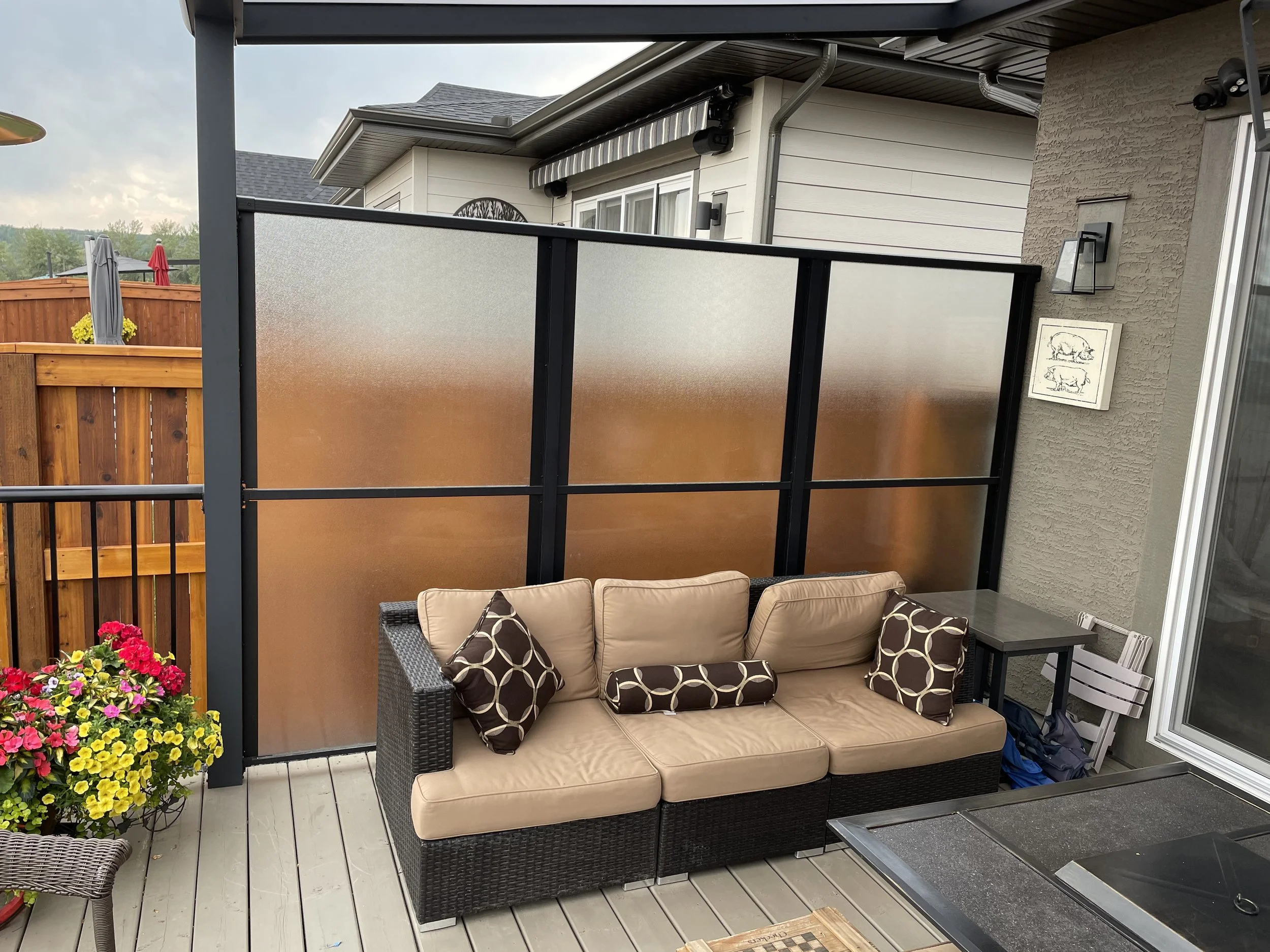 A cozy outdoor patio area with a beige cushioned wicker sofa, patterned pillows, a small side table, and a colorful potted flower arrangement, enclosed by frosted glass privacy screens and wooden fencing, with a house wall, porch ceiling, and outdoor
