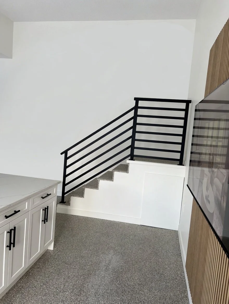 Interior of a modern home with a staircase featuring black metal railing, white walls, a white cabinet with black handles, and a large TV mounted on a wall with vertical wooden paneling.