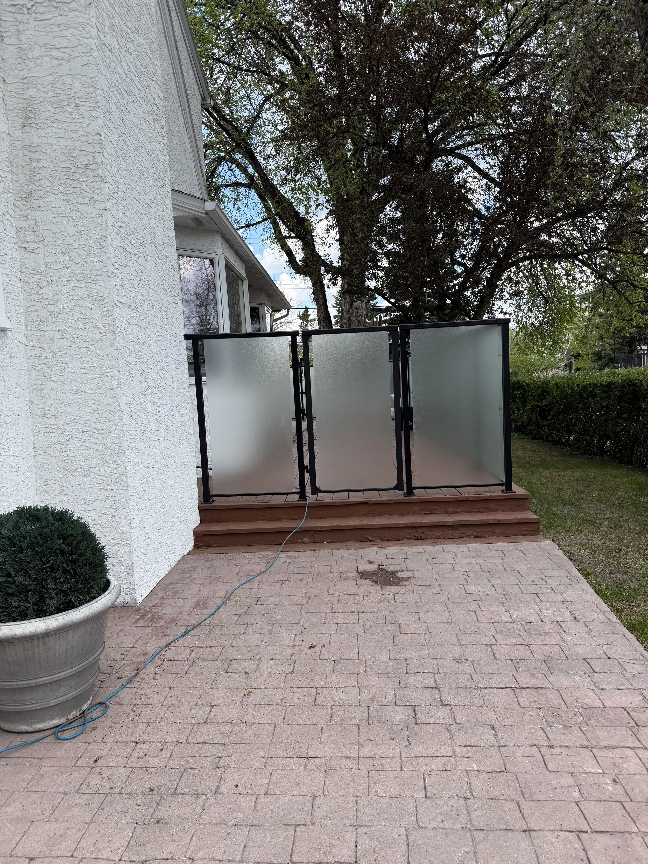A backyard patio with red brick pavers, a white textured house wall on the left, a potted plant in the corner, a black metal patio railing with frosted glass panels, a large tree with green leaves and brown branches, and a grassy yard with bushes in 
