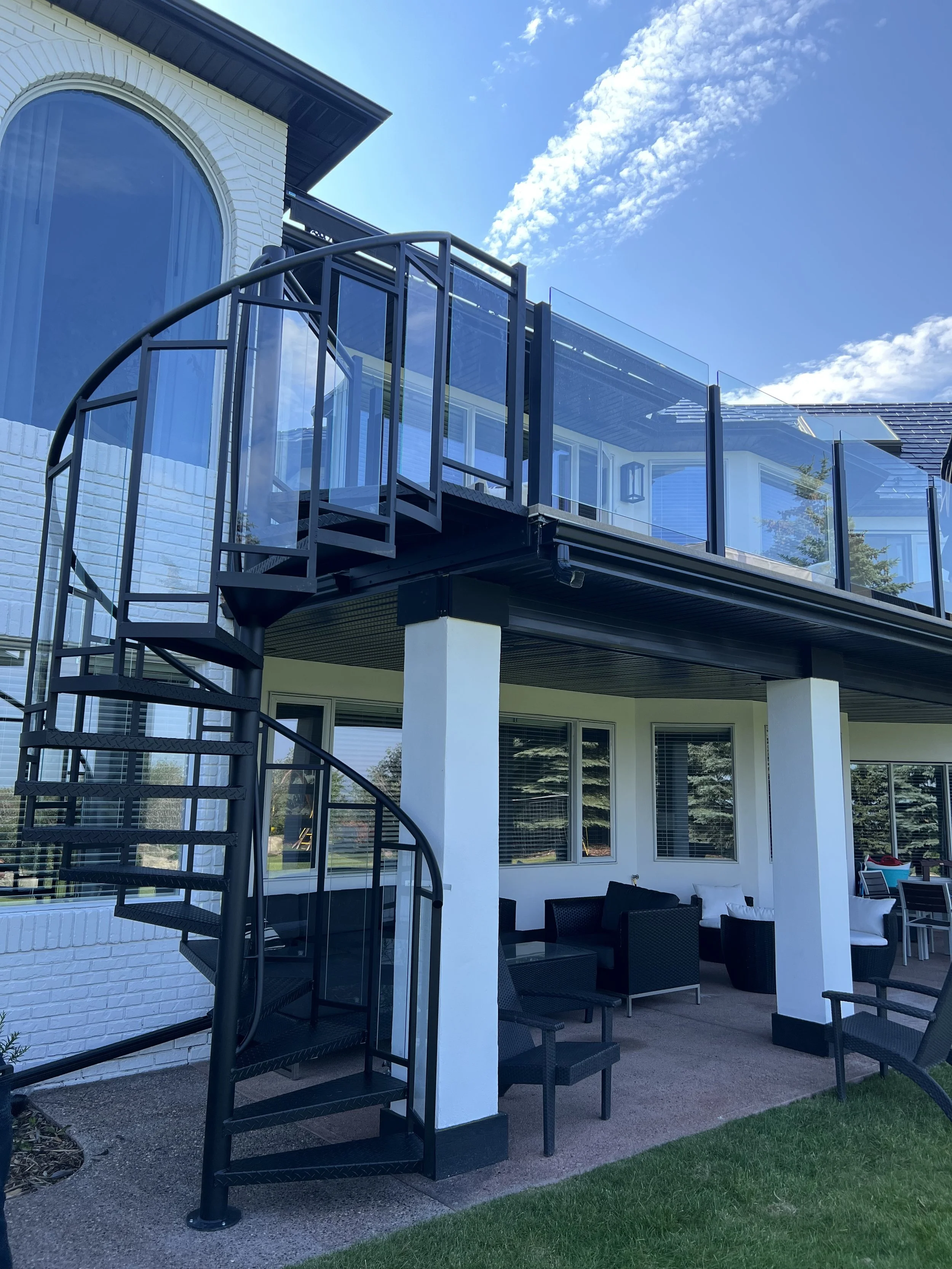 Backyard patio area with outdoor seating, a spiral staircase leading to an upper balcony, and large glass windows reflecting the sky.