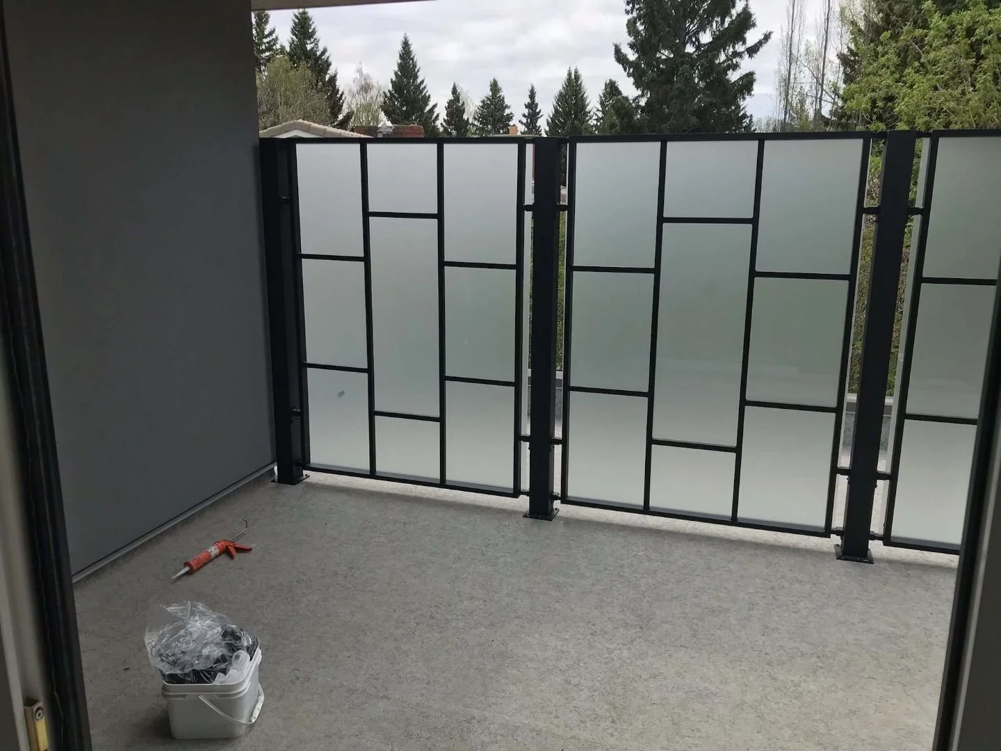 Balcony with frosted glass and black metal railing, overcast sky, trees in background, construction tools and supplies on carpeted floor.