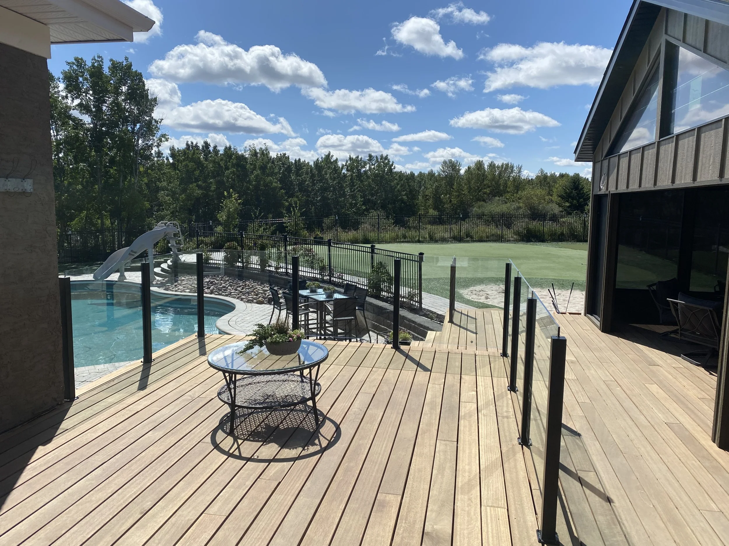 View of a backyard patio with a swimming pool, outdoor furniture, and a fenced grassy area with trees in the background. The sky is blue with scattered clouds.
