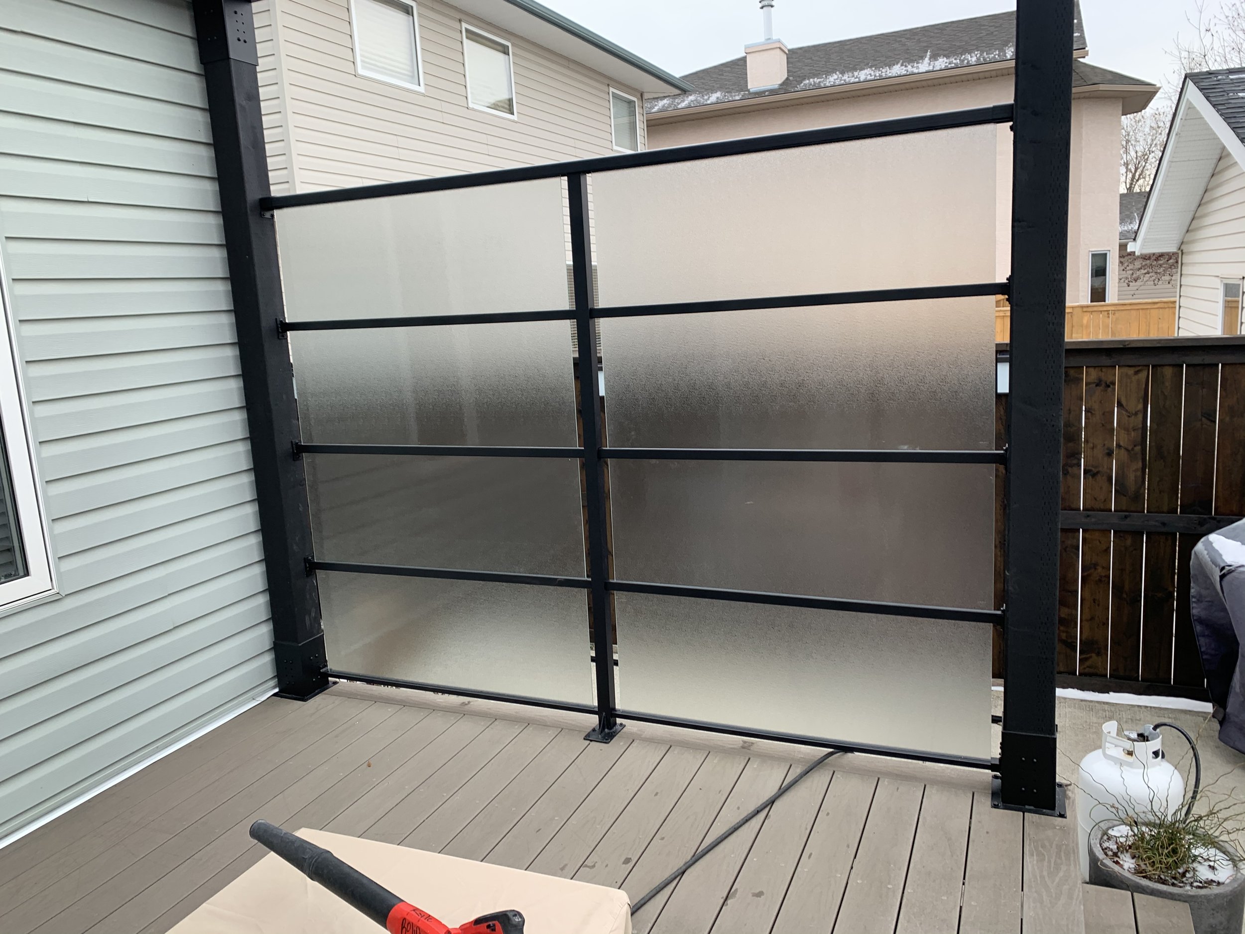 A partially assembled outdoor deck railing on a wooden deck, with a black metal frame and frosted glass panels, next to a house with beige siding.