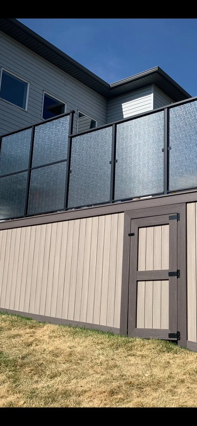 Modern house exterior showing siding, windows, a balcony with textured glass panels, and a small gate on a grassy area.