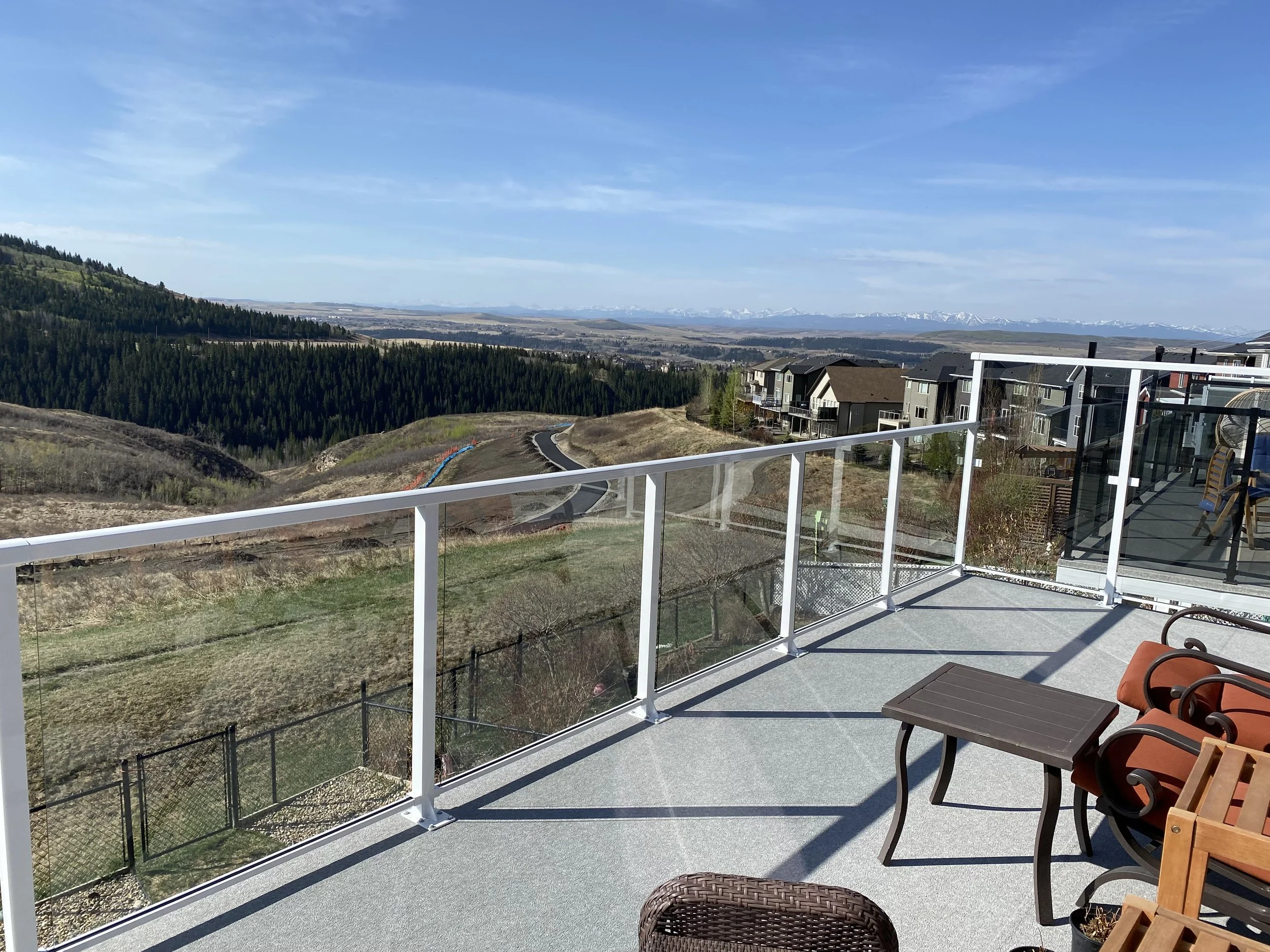 A balcony with orange and brown chairs, overlooking a natural landscape with mountains in the distance and houses below, under a blue sky with some clouds.