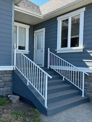 Front entrance of a house with gray stairs, white railings, a blue exterior, and a white door surrounded by two large windows.