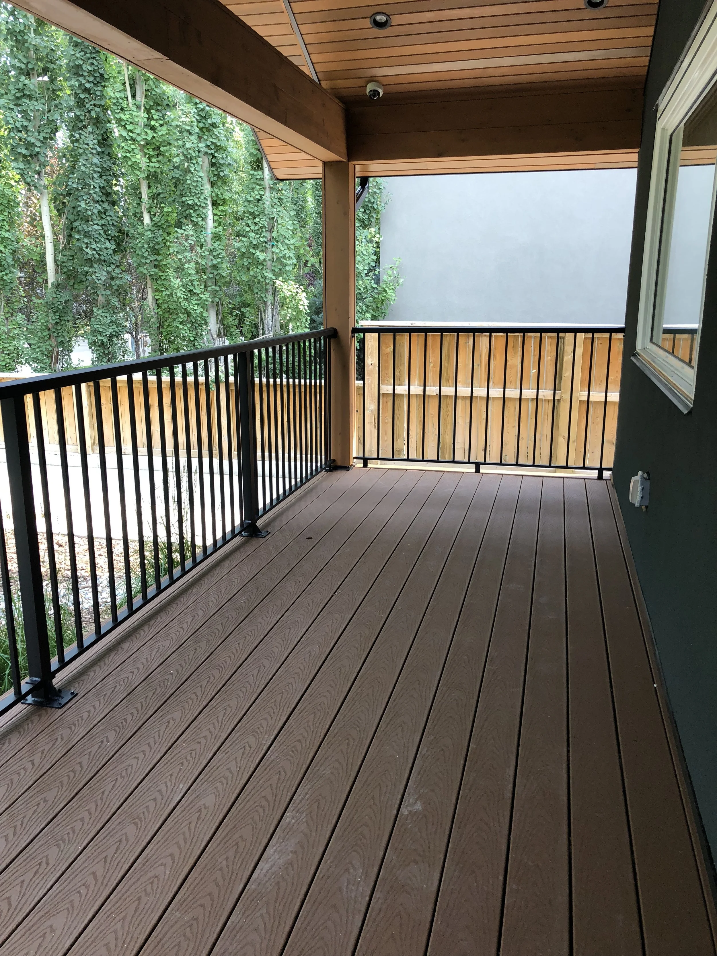 A wooden balcony with a black metal railing, overlooking trees. The ceiling and the floor are made of matching warm-toned wood. There is a security camera and a smoke detector on the ceiling, and a white electrical outlet on the wall.