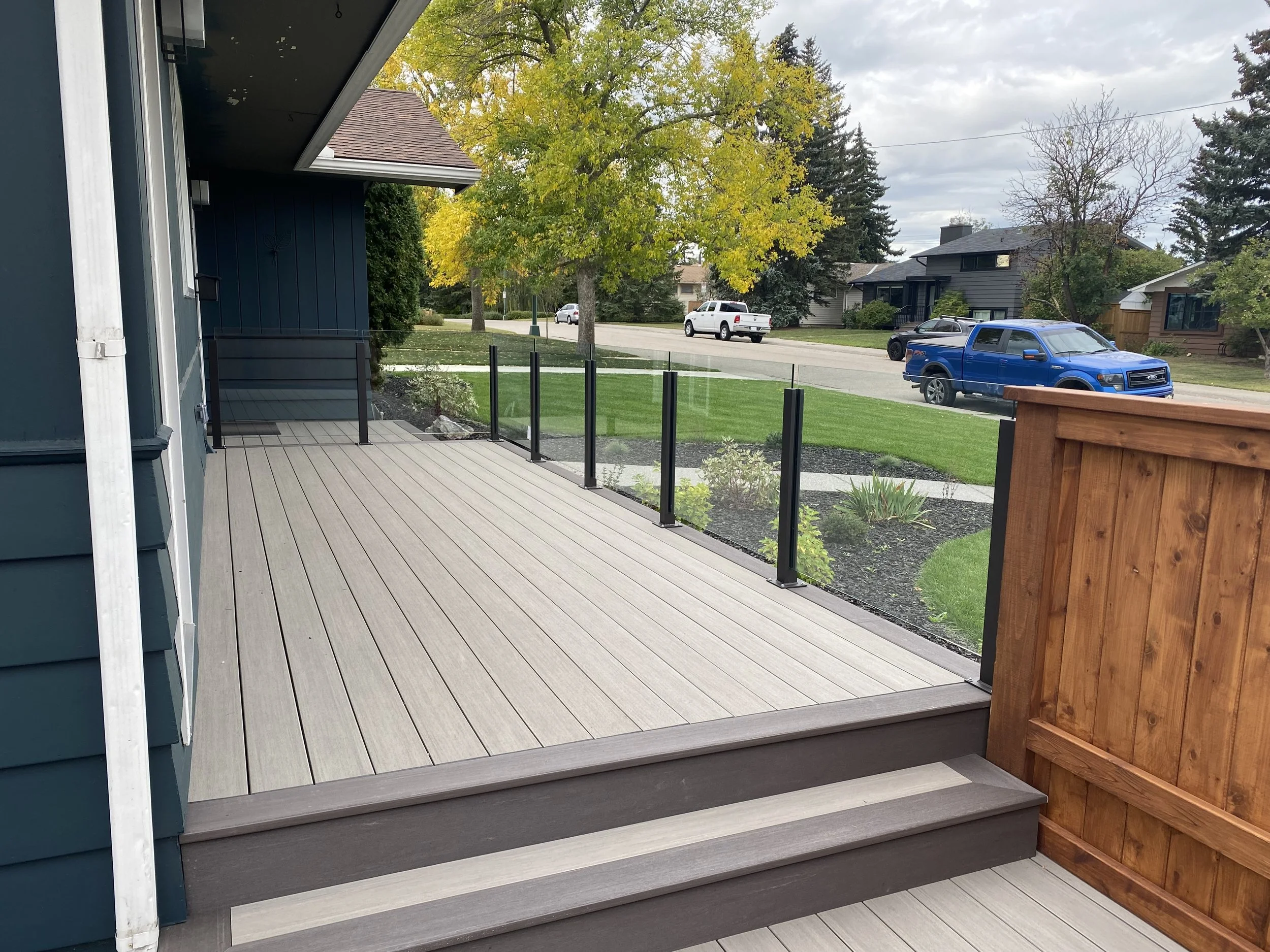 View of a neighborhood street seen from a backyard wooden deck with a glass railing. The street has parked cars and houses with trees showing fall foliage, some leaves turning yellow and brown. The sky is cloudy.