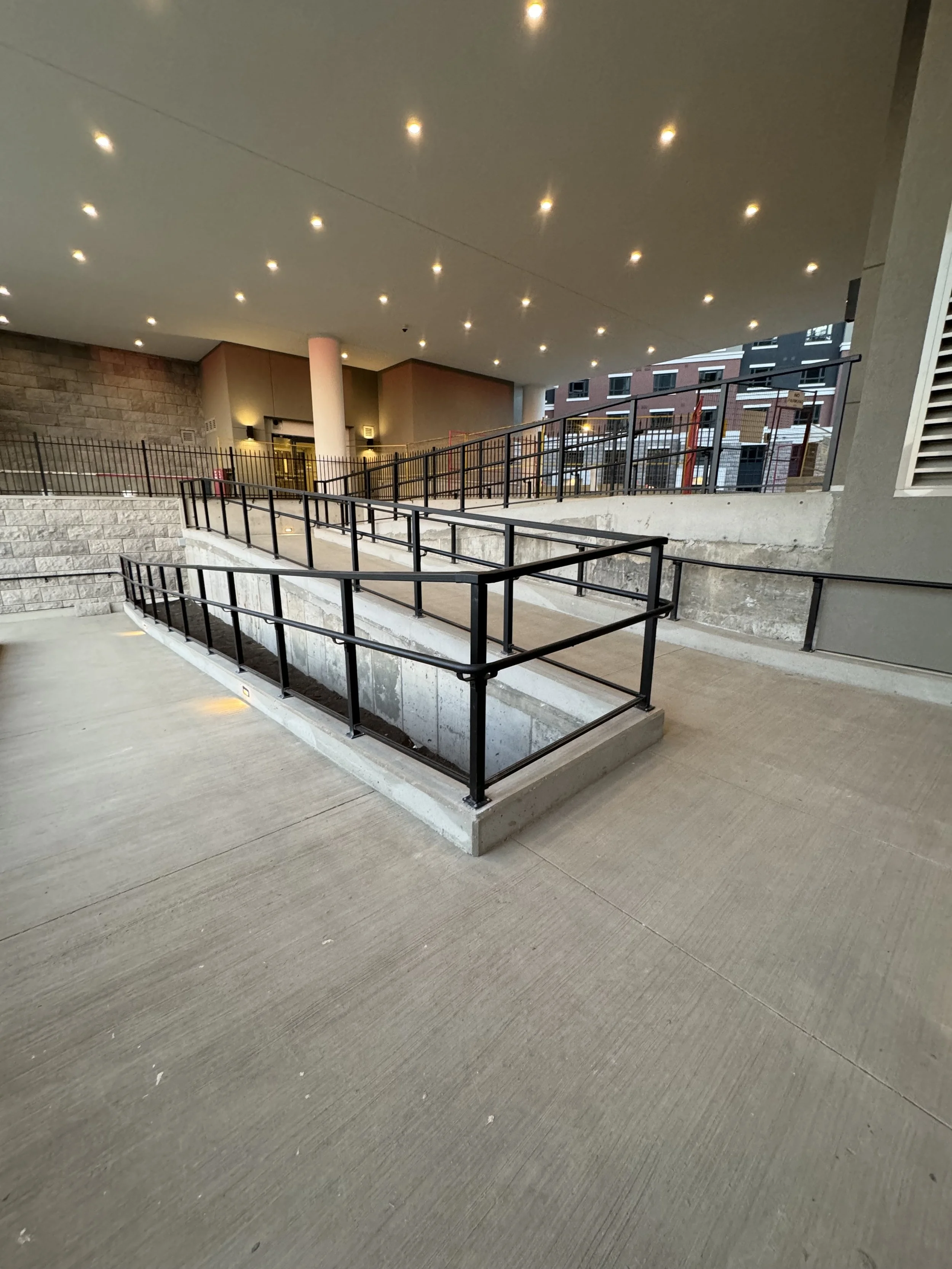 A multi-level parking garage with concrete floors, black metal railings, and a ceiling with many small bright lights; part of a cityscape with residential buildings visible in the background.