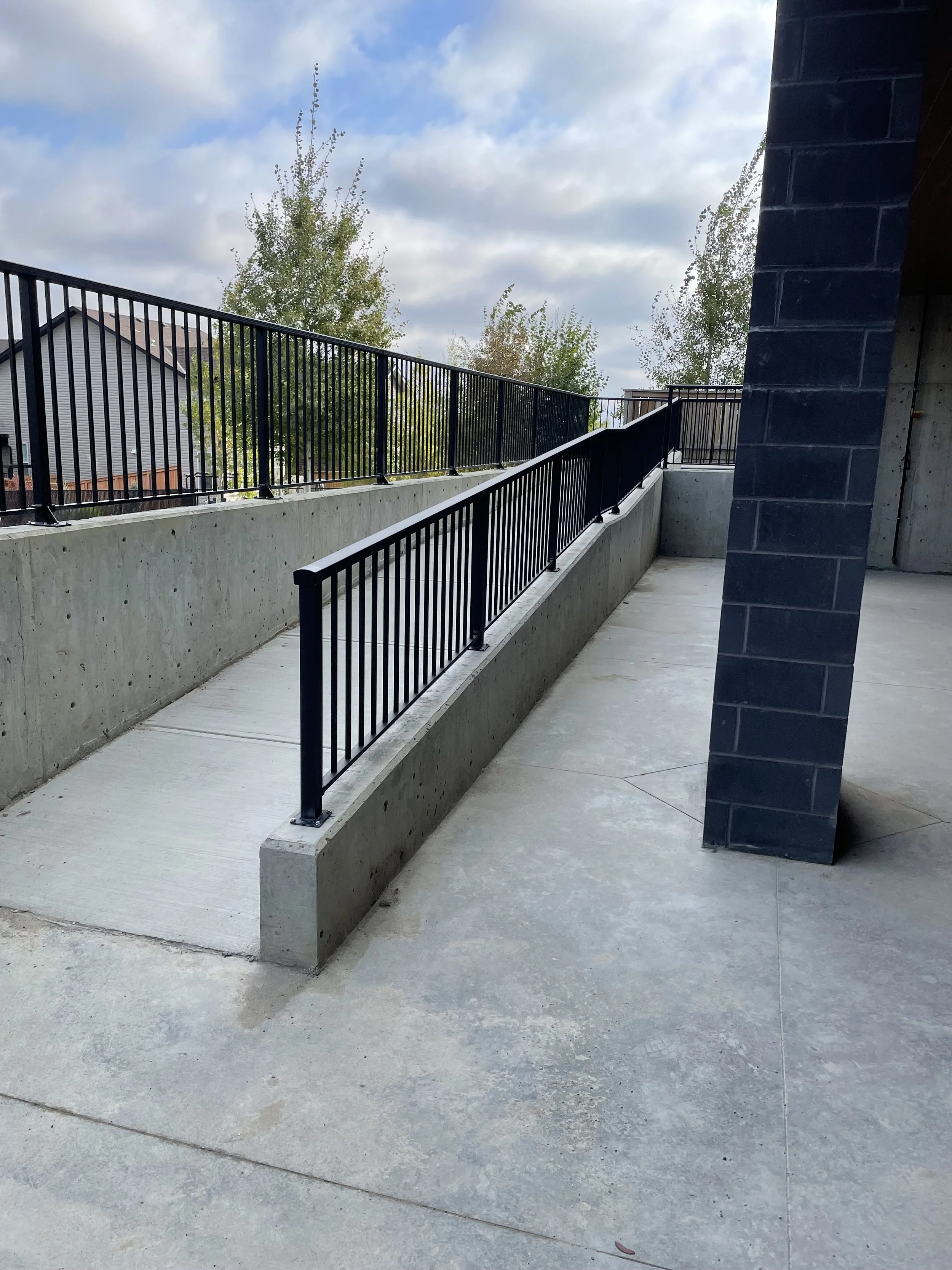 An outdoor balcony or walkway with a concrete floor and a black metal railing. Part of a building wall made of dark bricks is visible on the right. In the background, there are trees and houses under a partly cloudy sky.