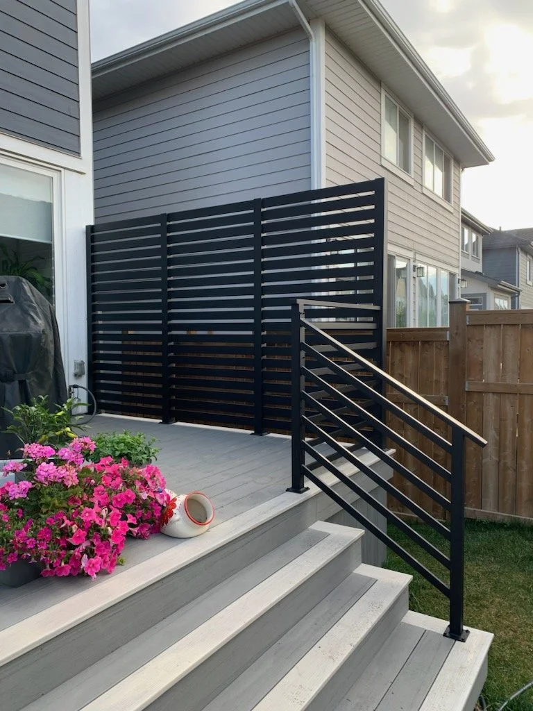 Backyard patio with gray steps, pink flowers, a black railing and privacy fence, with a house in the background.