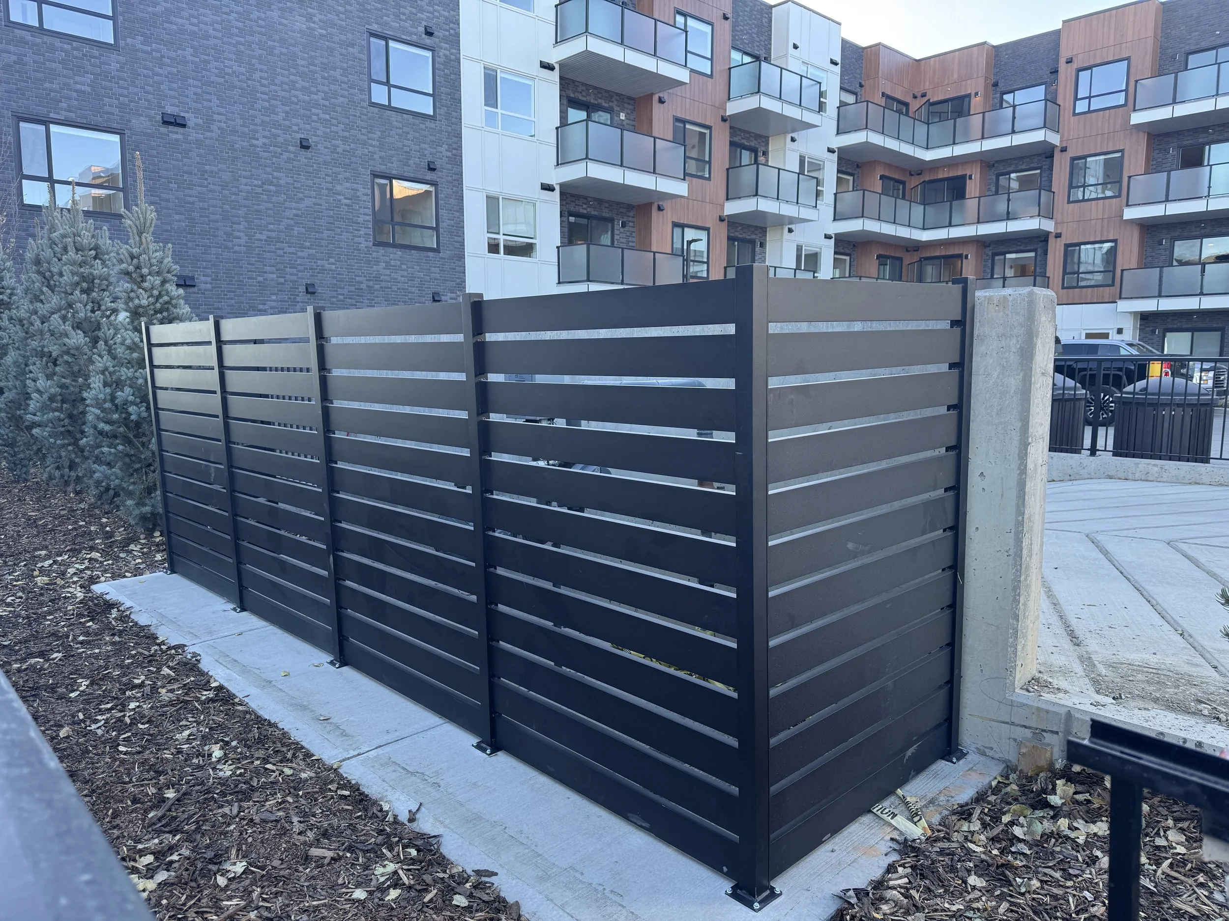 Modern apartment building with balconies, a black wooden fence, and a small landscaped area with a bush, situated in an urban setting.