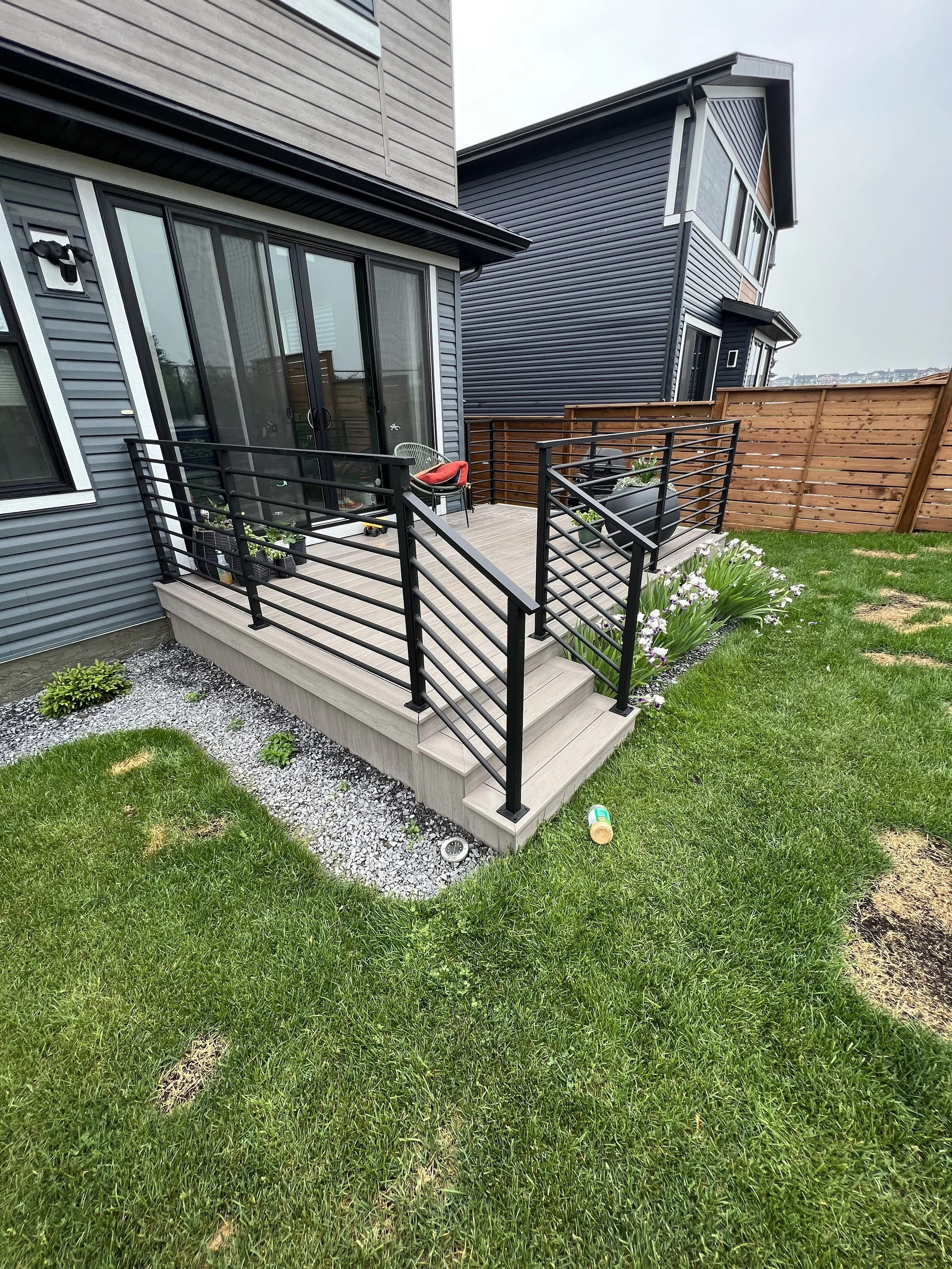 A small backyard patio with a wooden deck, black metal railings, potted plants, a chair with a red cushion, and a sliding glass door leading indoors. The yard has green grass, some flowers, and a wooden privacy fence.