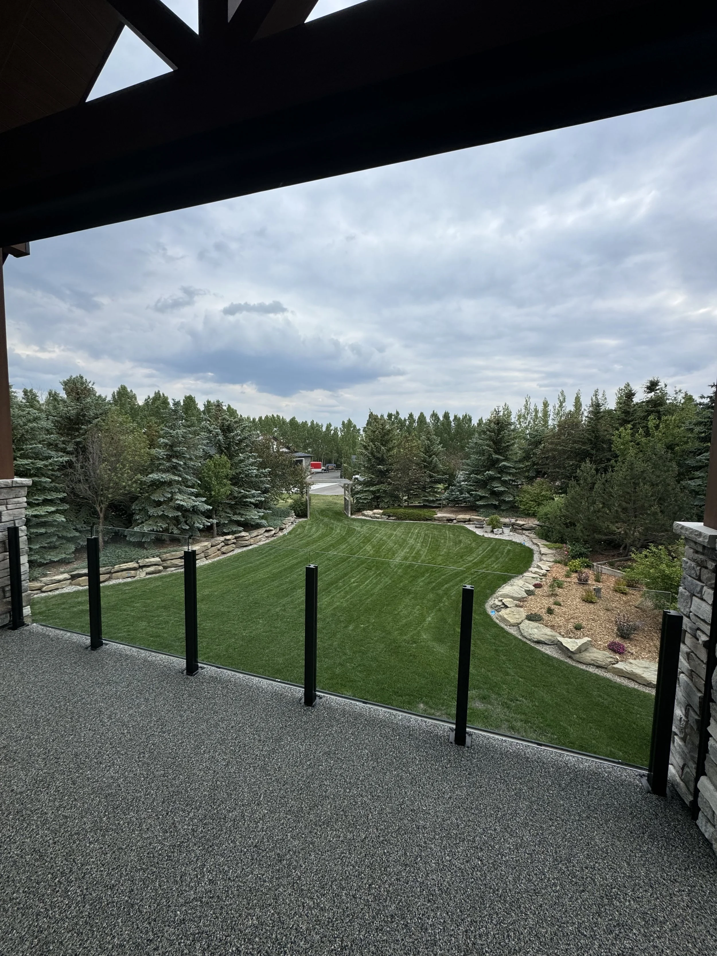 View from a balcony with a black rope railing, overlooking a yard with a green lawn, bordered by rocks and trees, under a cloudy sky.