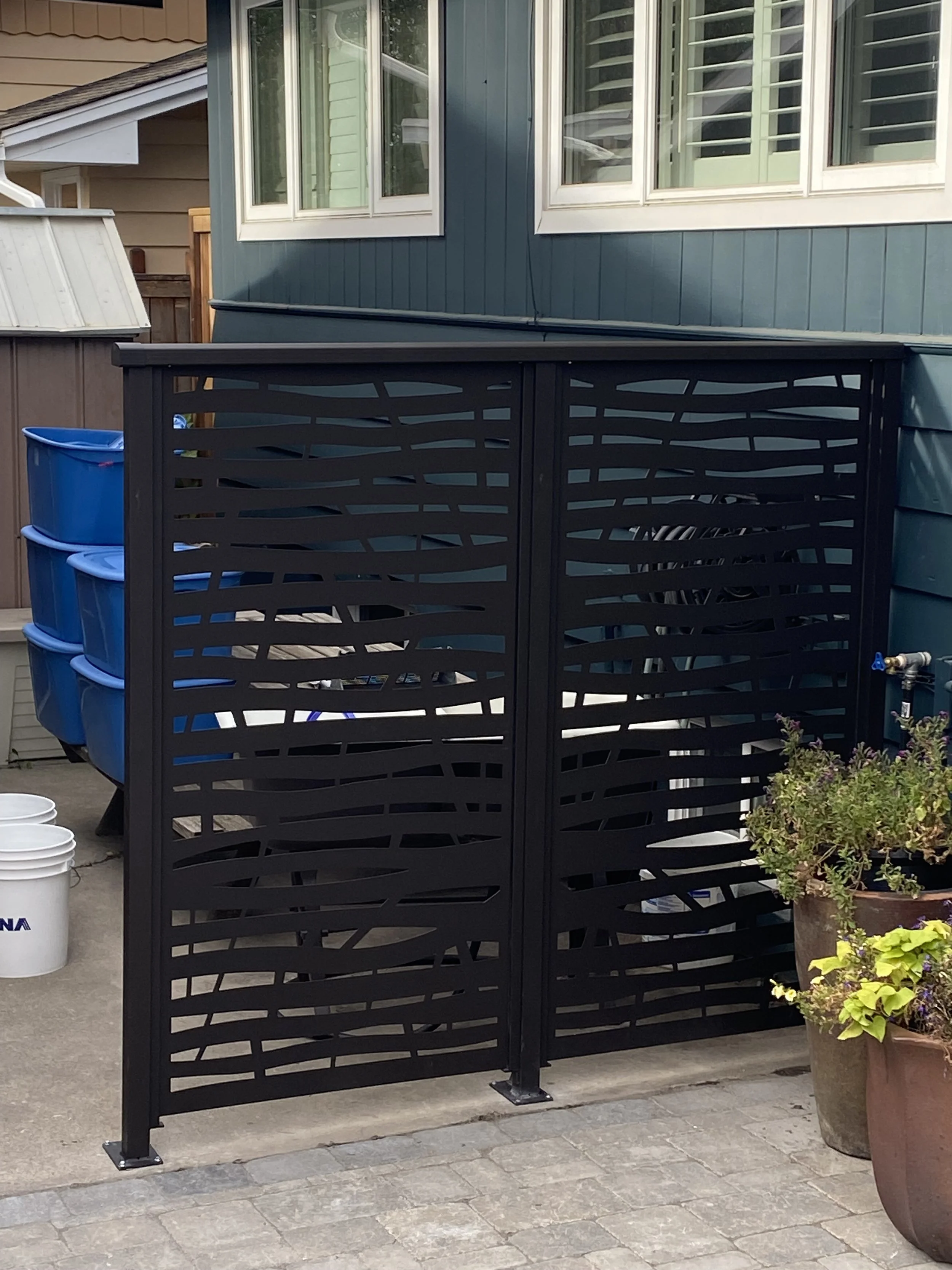 A black metal gate with a wavy cutout design, installed on a paved outdoor space, adjacent to a large potted plant with green and purple foliage, and blue storage containers stacked on the left side of the image. In the background, there is a house w