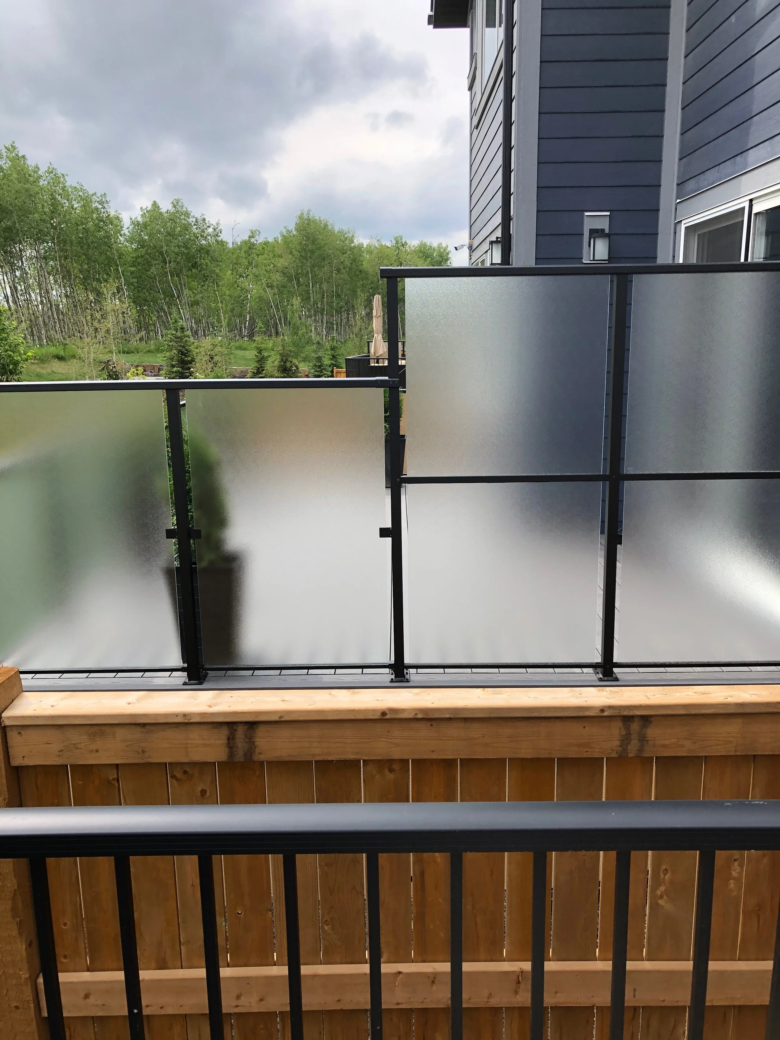 View from a balcony showing a frosted glass railing with black metal framing, a blue house exterior, and a wooden deck below. In the background, there are green trees and gray storm clouds in the sky.