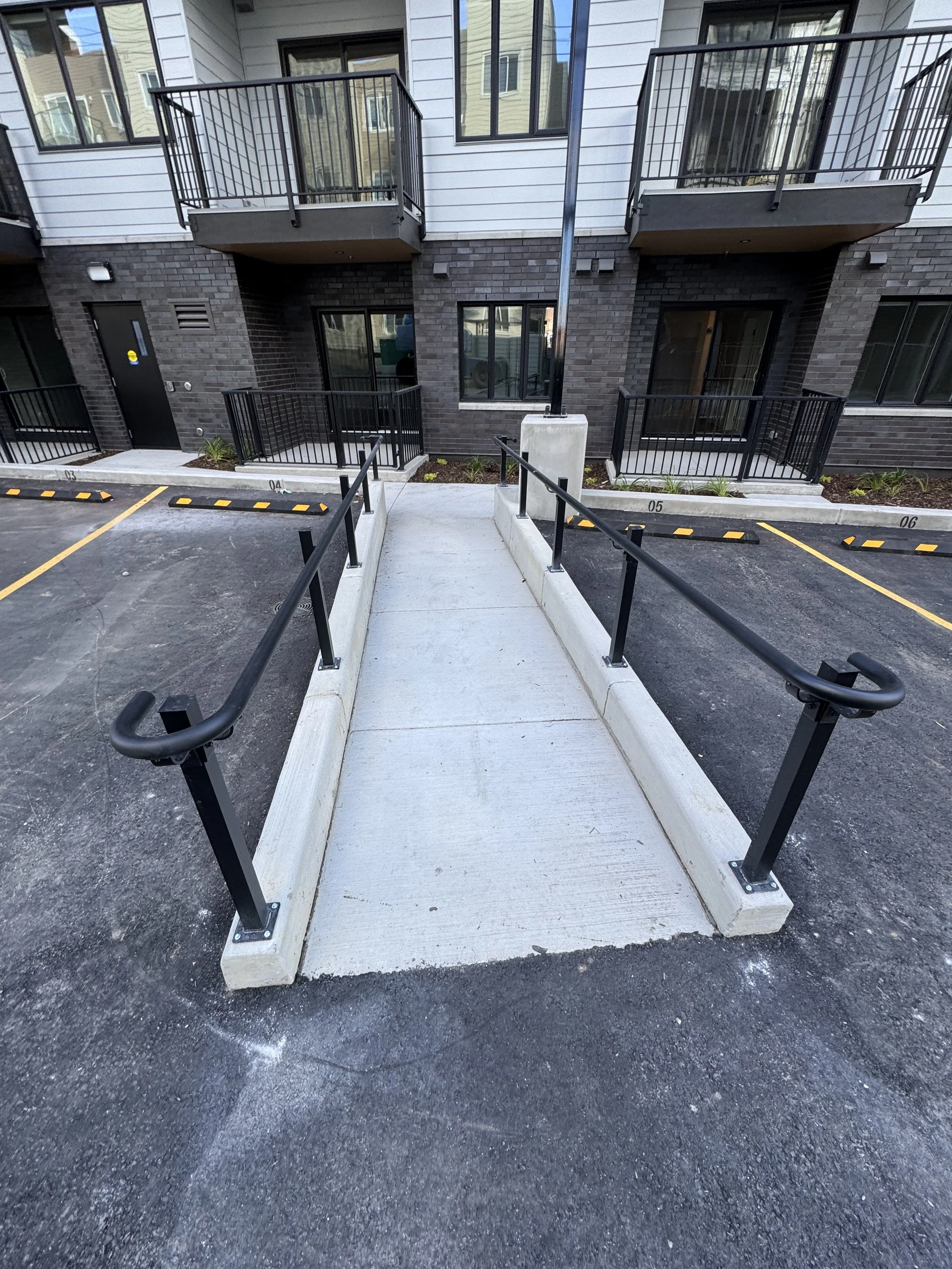 A wheelchair accessible ramp leading to the entrance of an apartment building with parking spaces in front.