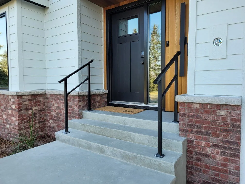 Front entrance of a house with a black door, concrete steps, and black metal handrails, surrounded by white siding, brick accents, and wooden trim.