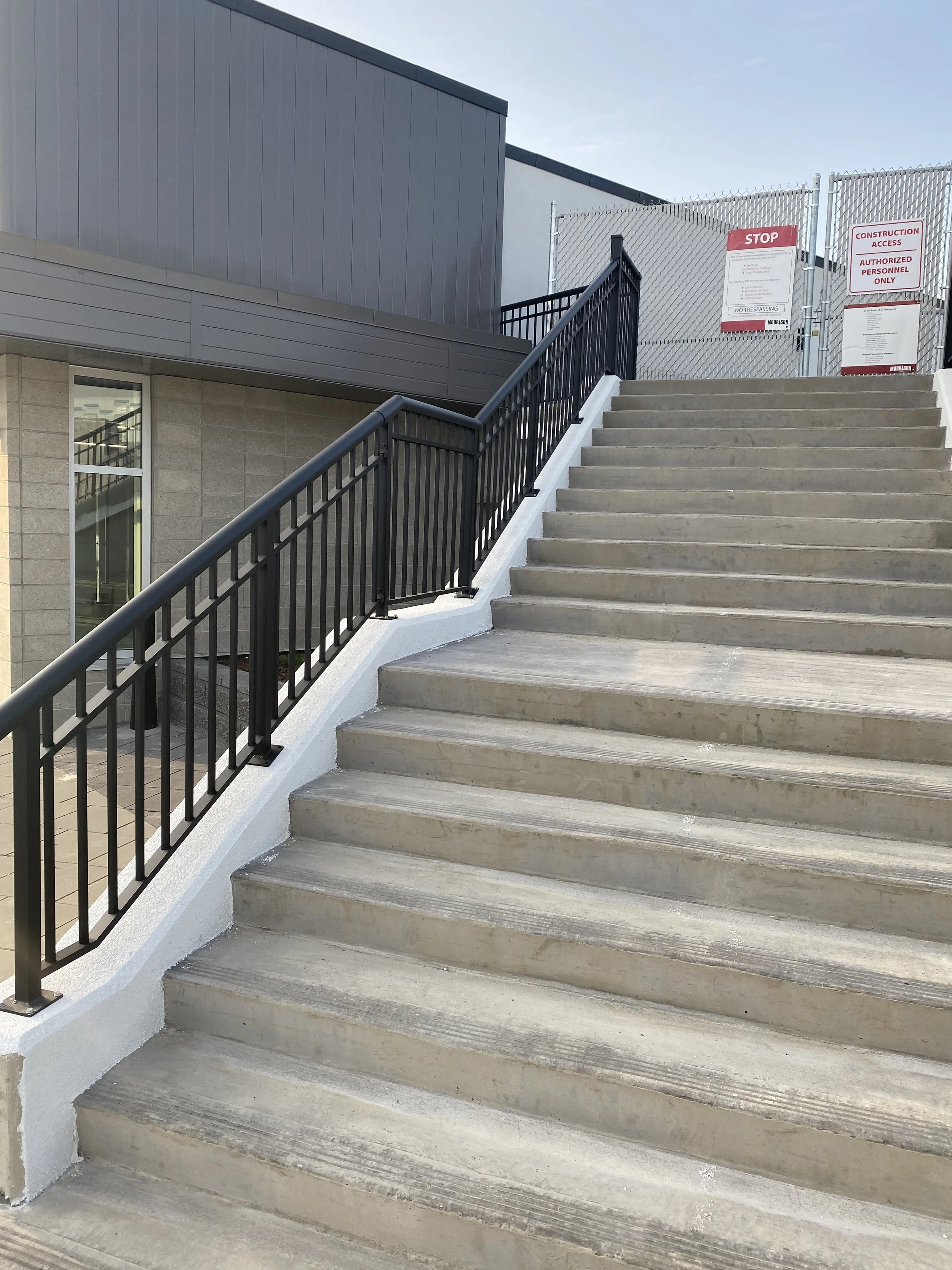 Outdoor concrete staircase with black metal handrails leading up to a fenced area with construction signs and partial view of a building.