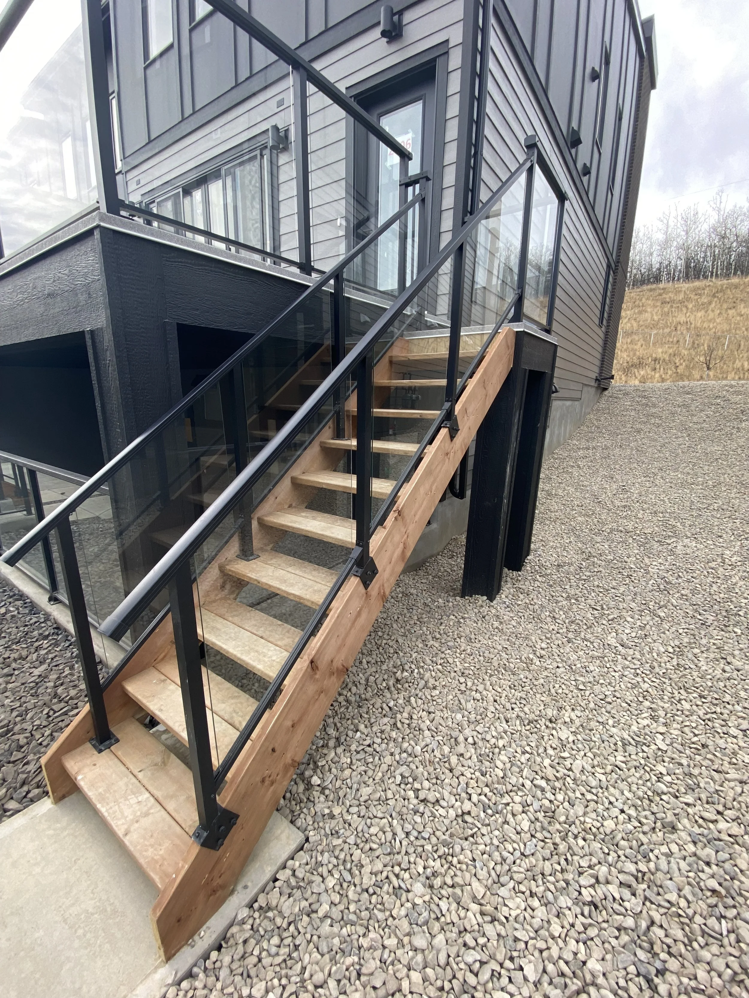 Exterior of a modern house with an outdoor staircase made of wood and metal, leading to a glass door with a window, with gravel landscaping surrounding it.