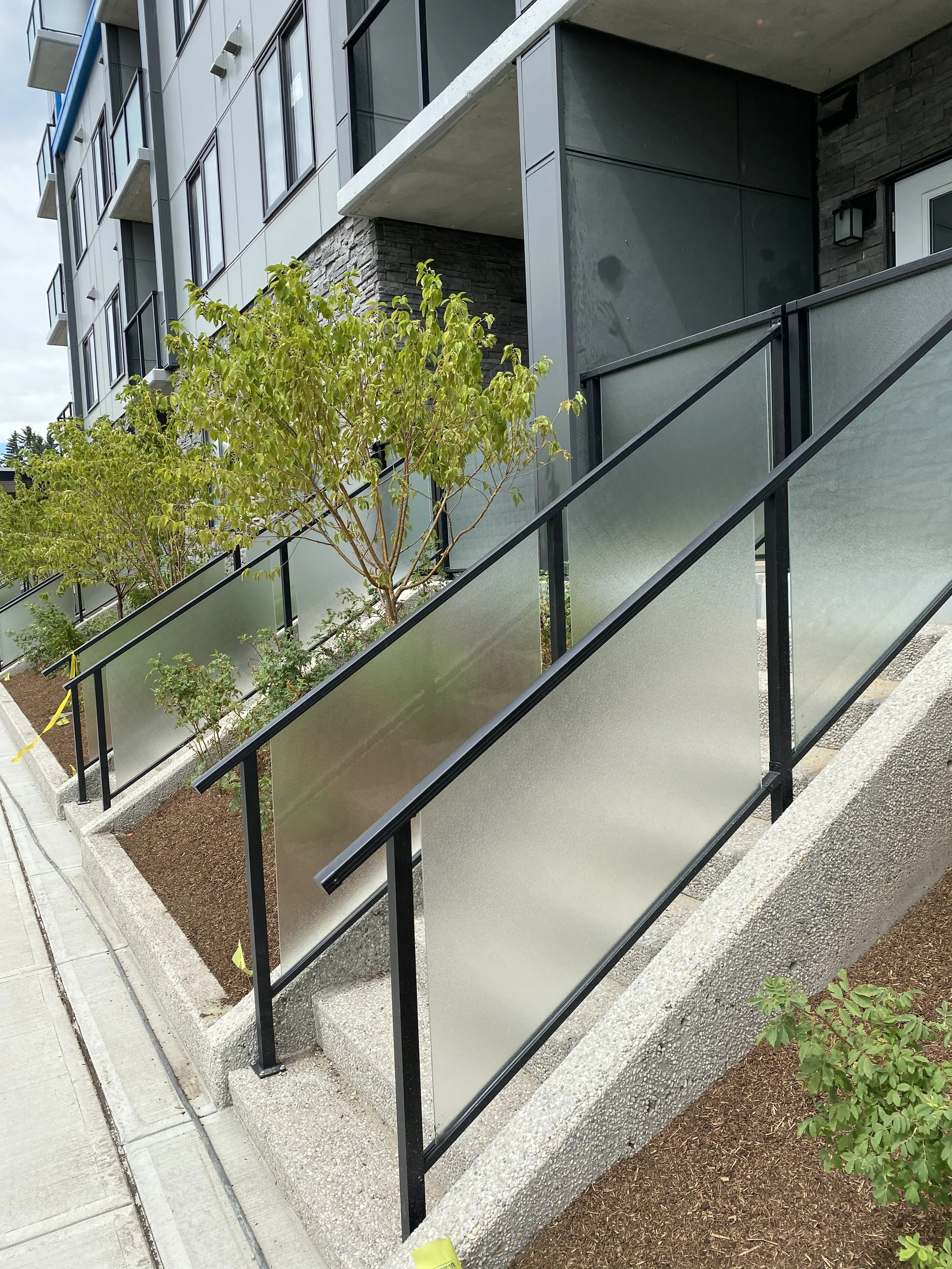Modern apartment building with outdoor ramp, frosted glass panels, black metal railings, brick and concrete exterior, and trees in landscaped area.