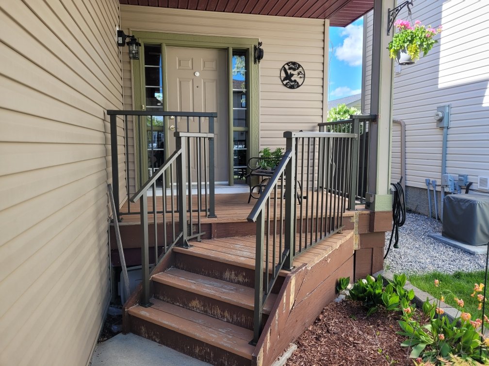 Front porch of a house with beige siding, brown steps, black and beige railing, potted plant, and a decorative wall hanging with a tree silhouette. There are plants and flowers in the garden soil below the steps and a hanging flower basket on the por