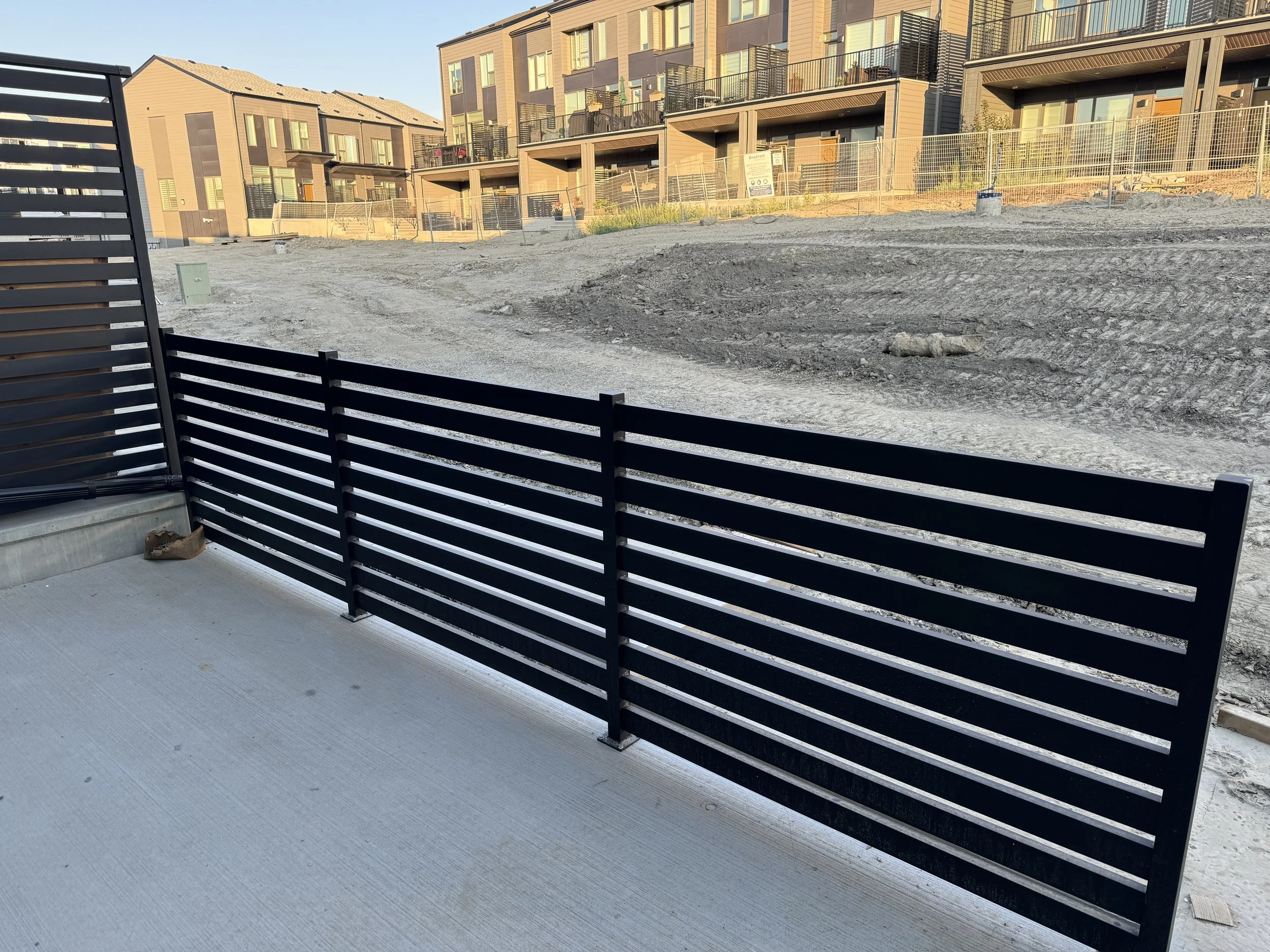 View of an outdoor construction site with a black wooden fence, dirt ground, and a modern multi-story residential building in the background.