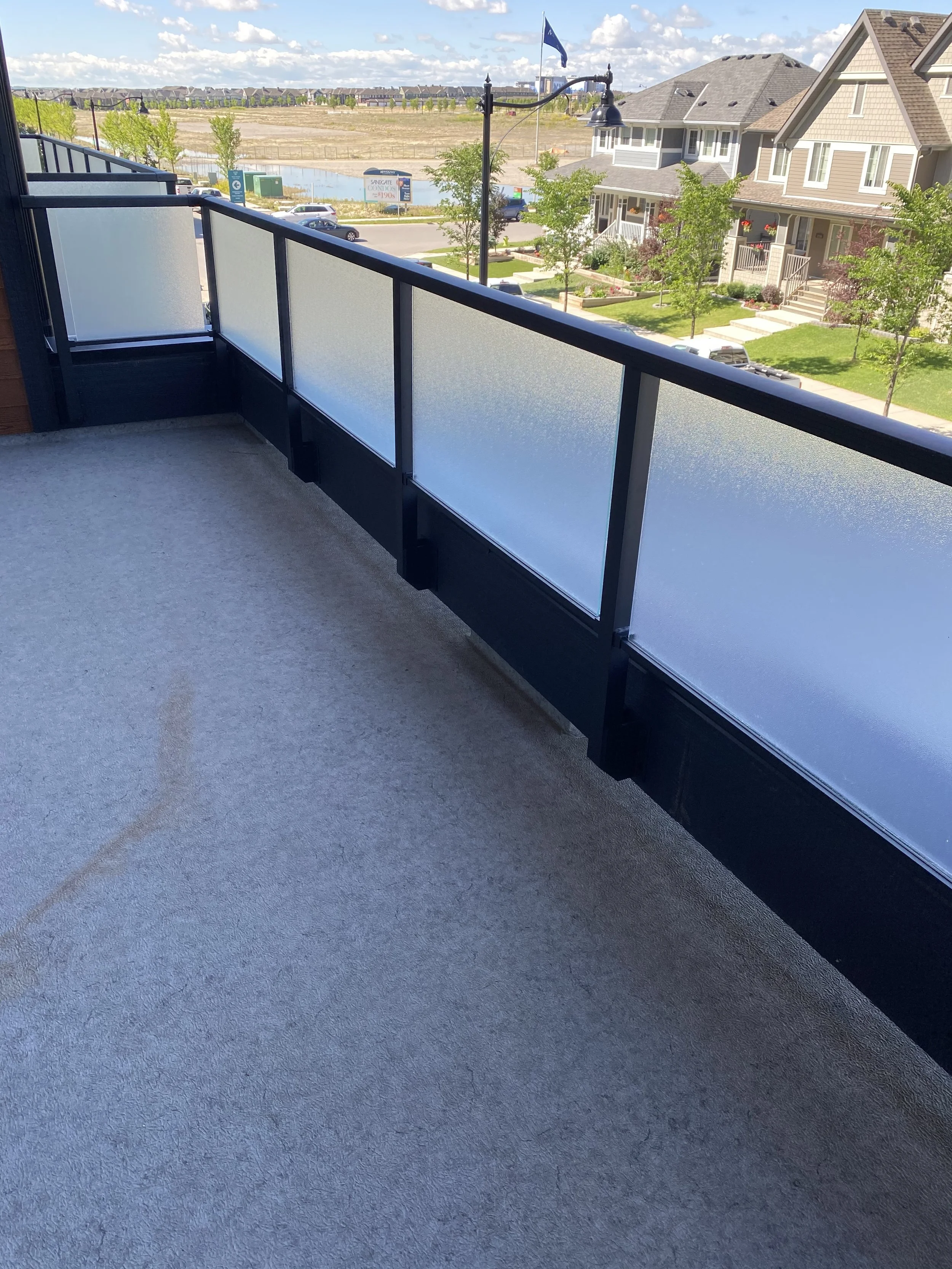 A balcony with a black metal railing featuring frosted glass panels, overlooking a suburban neighborhood with houses, trees, a street, parked cars, and a large open field in the background under a partly cloudy sky.