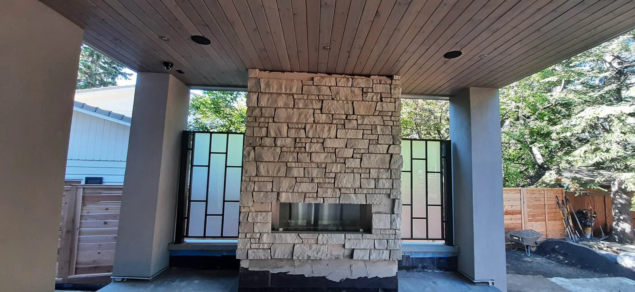 Outdoor living space with a stone fireplace, wooden ceiling, and partial view of a backyard with trees and fencing.