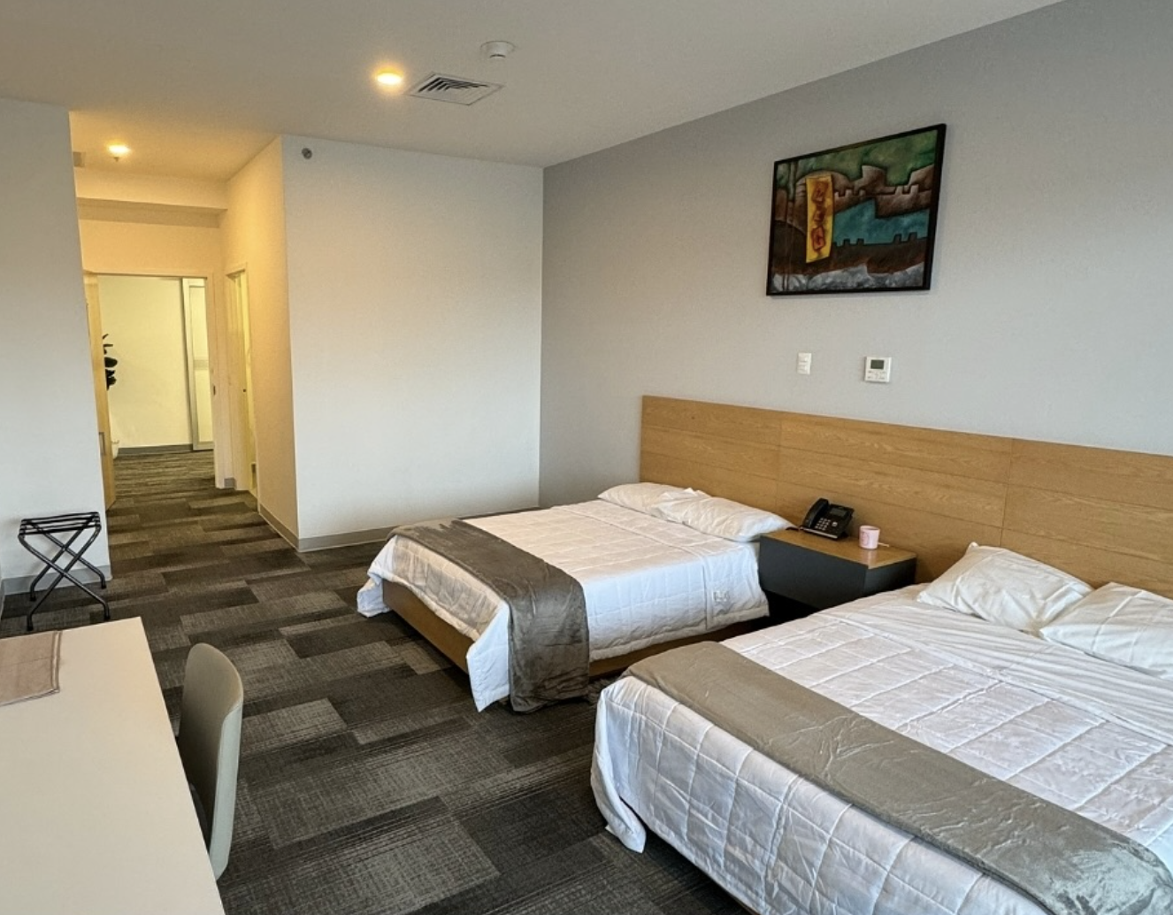 Hotel room with two beds, a nightstand with a phone and pink candle, artwork on wall, doorway to hallway, and patterned carpet.