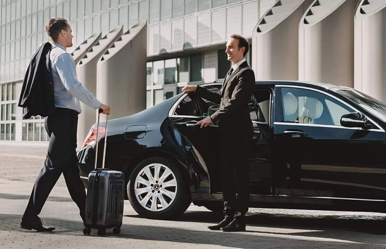 A man in a suit standing next to his car and talking to a traveler with a suitcase outside a modern building.