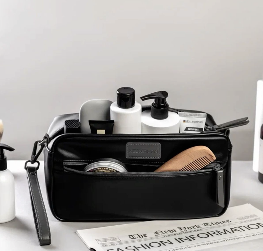 A black toiletry bag filled with various bottles, a hairbrush, and grooming products on a white surface, with a newspaper partially visible in front.