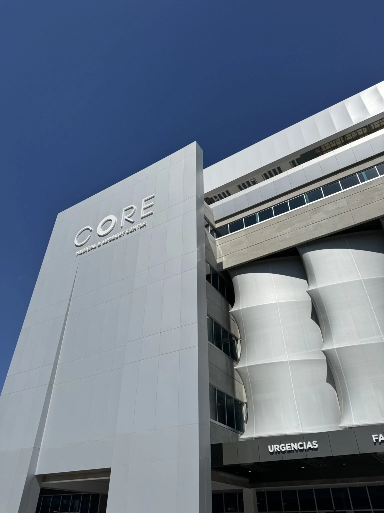 Modern hospital building with white exterior, displaying the words 'CÓRRE' and 'URGENCIAS' over the entrance, against a clear blue sky.