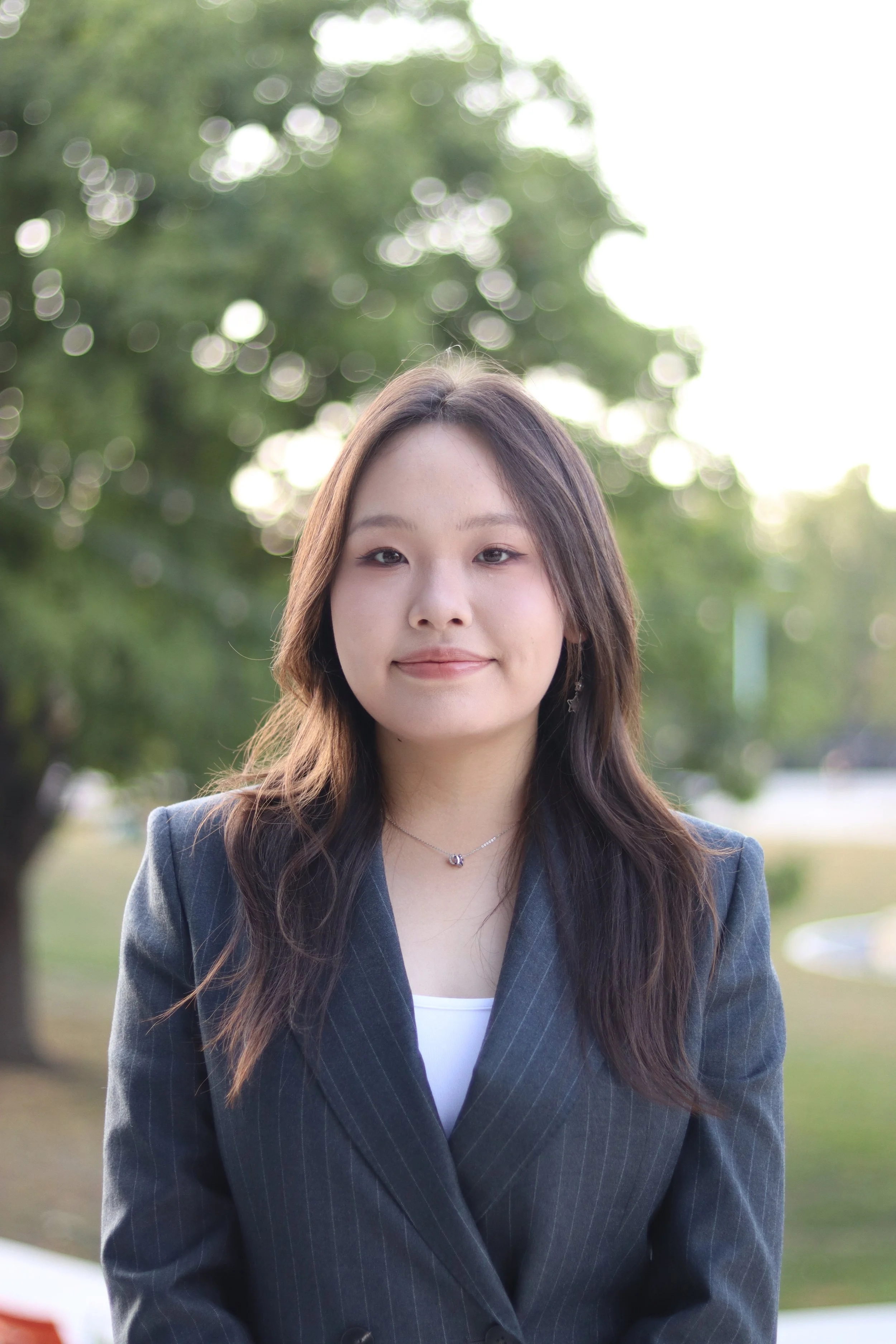 A young woman with long wavy brown hair wearing a dark pinstripe blazer over a white top standing outdoors with trees and bright sunlight in the background.