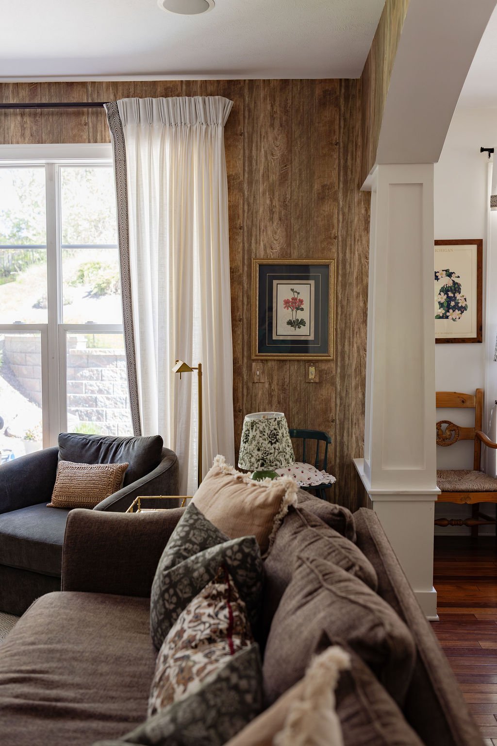 Living room corner with sofas, decorative pillows, a small lamp, and wall art, near a window with white curtains and wooden wall paneling.