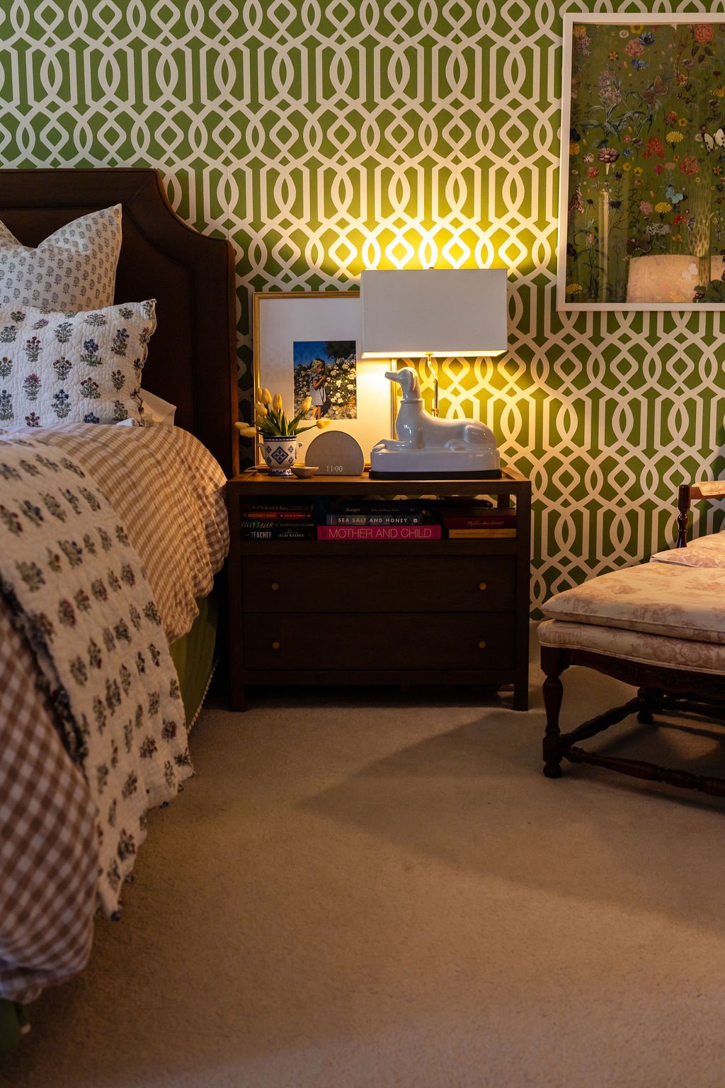 A bedroom with a dark wood bed, floral patterned bedding, a nightstand with books, a lamp, a decorative dog-shaped humidifier, a framed photo, and a wall with green geometric wallpaper. There is also a floral framed picture and a vintage-style cushio