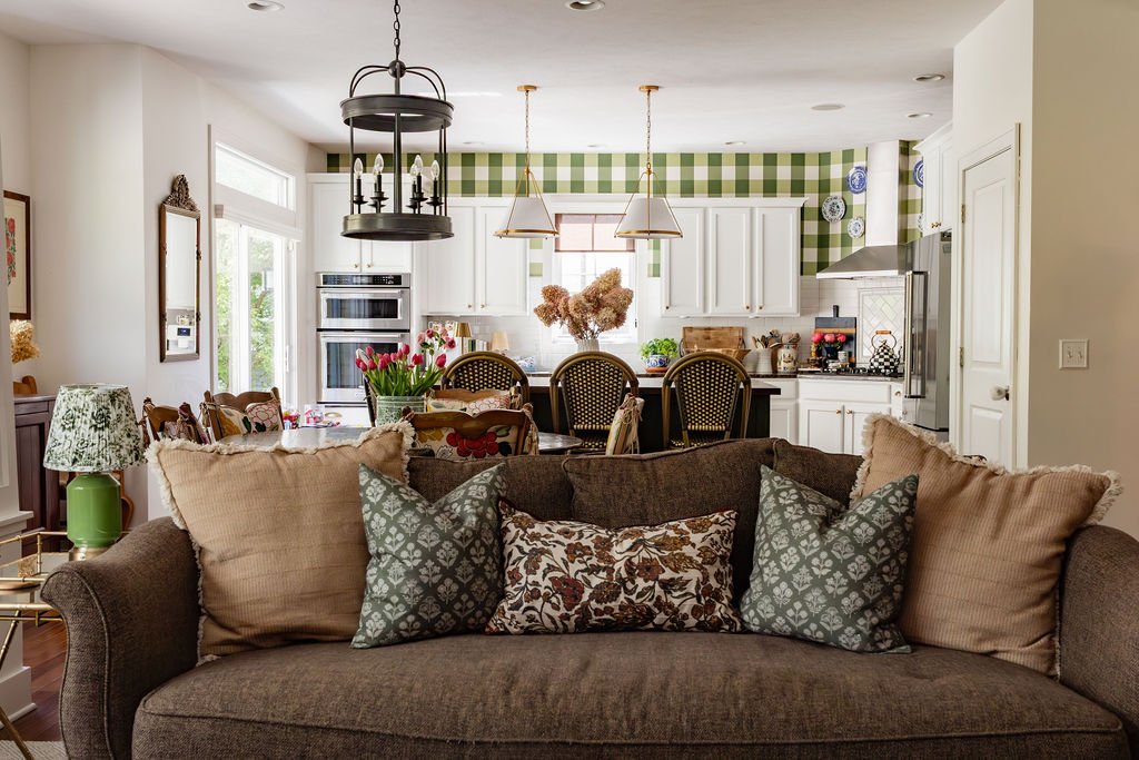 Living room with a brown couch and multiple decorative pillows, open kitchen with white cabinets, a green and white checkered wall, and a dining area with a round wooden table and chairs in the background.