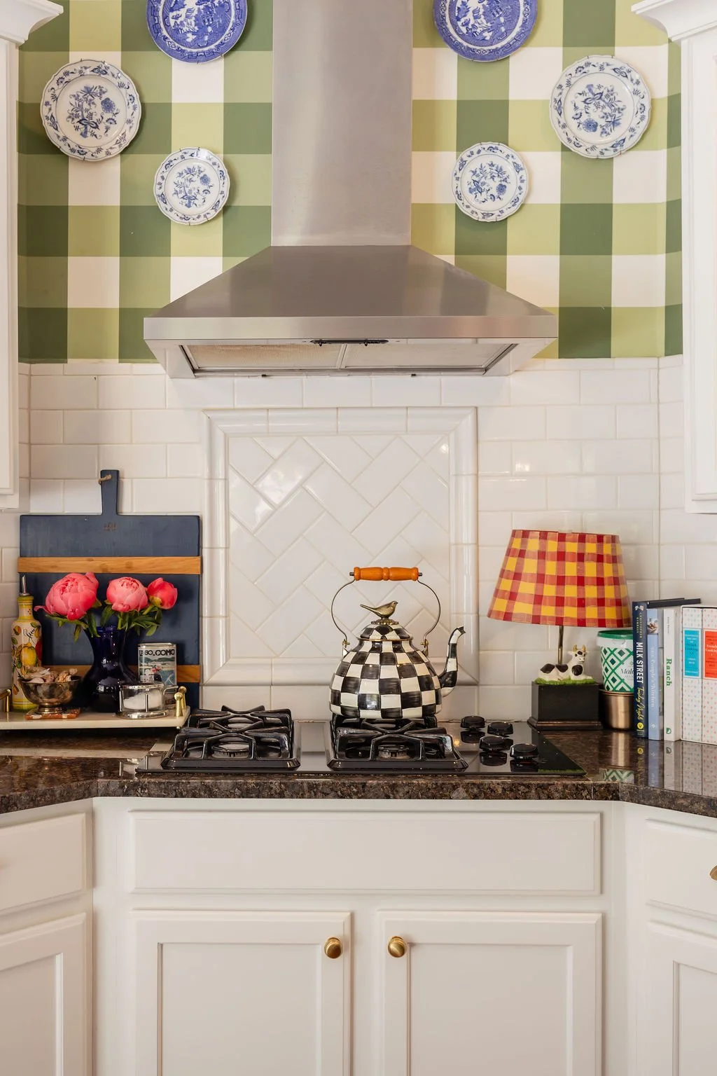 Kitchen with white cabinets, a black and white checkered kettle on a stovetop, a vase with pink flowers, a checkered lamp, and decorative plates on a green checkered backsplash.