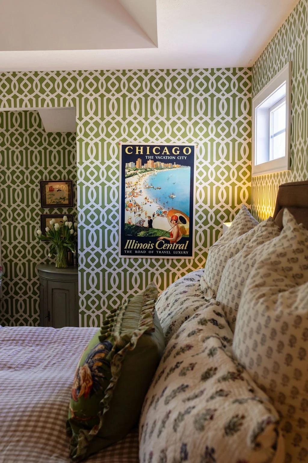 A bedroom with green and white geometric wallpaper, a bed with floral-patterned bedding, a poster of Chicago, a small bedside table with a vase of white tulips, and a small window.