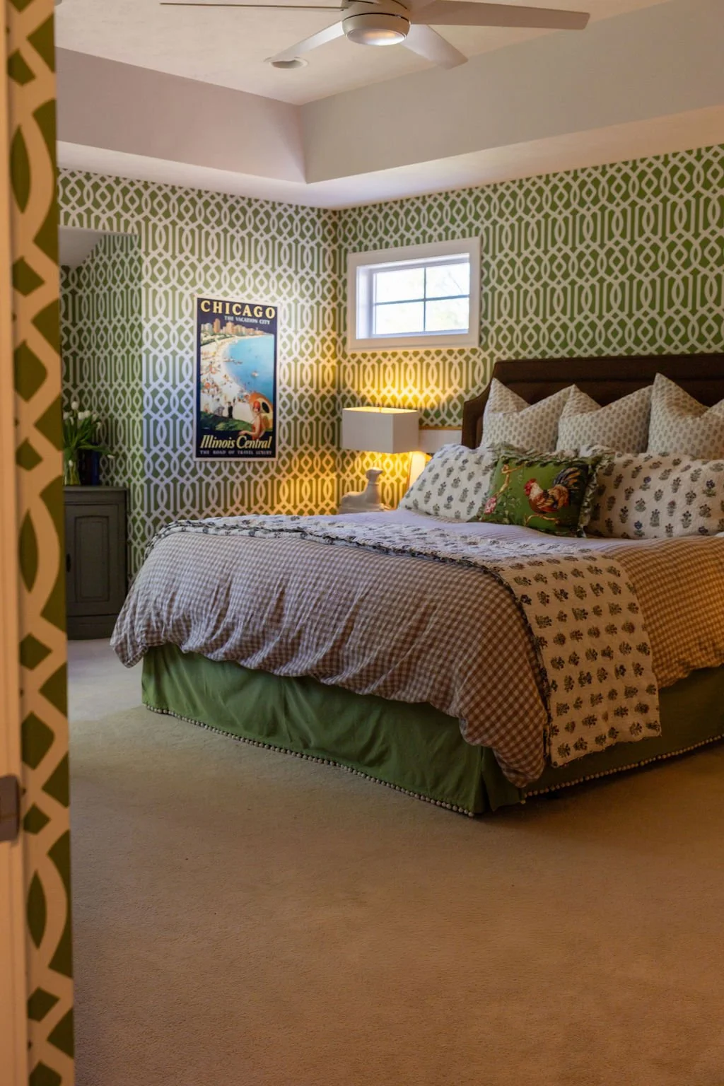 A cozy bedroom with a patterned wallpaper, a bed with multiple pillows and a decorative pillow featuring a rooster, a nightstand with a lamp, a window, and a vintage Chicago travel poster on the wall.