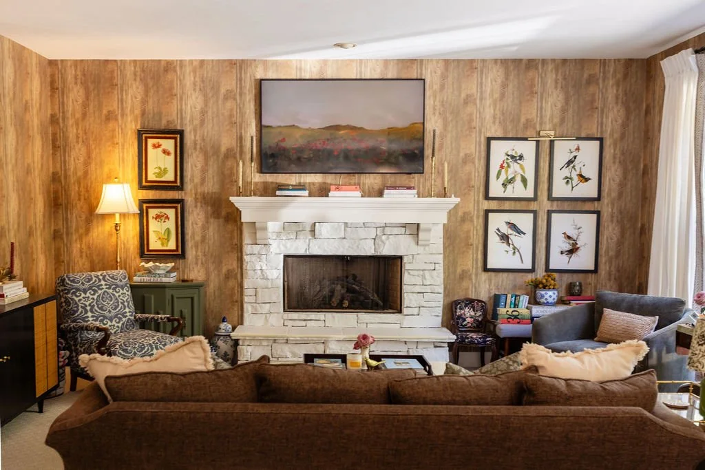 Living room with wood-paneled wall, central white brick fireplace, framed artwork including flowers and birds, a brown sofa, patterned armchair, and gray armchair, with a window and curtains on the right side.