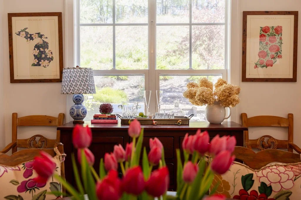A dining room with a window, floral artwork, a lamp, a vase with dried flowers, and tulips in the foreground.