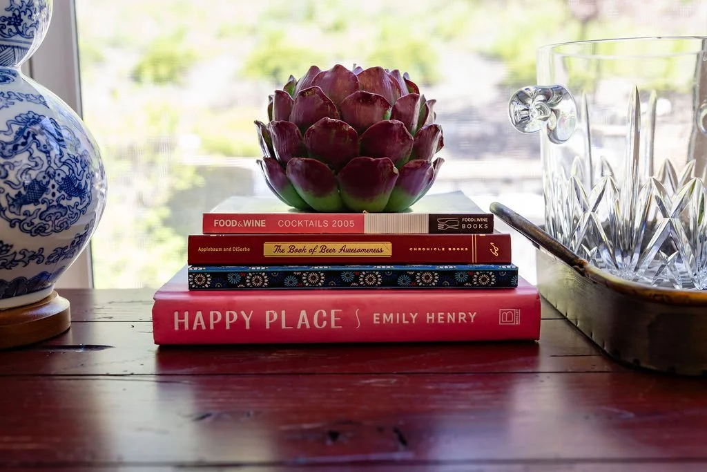 A stack of three books with a purple artichoke on top, placed on a wooden table beside a blue and white vase and a tray with glassware, with a window and greenery in the background.