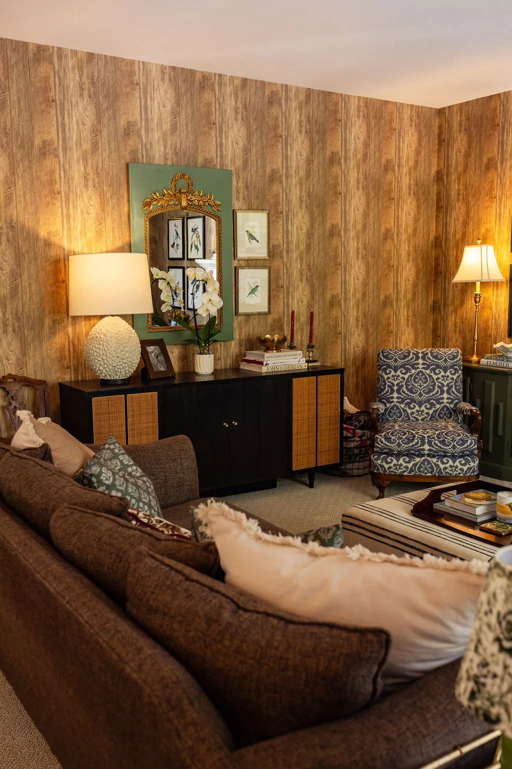 Living room with a brown sofa, patterned armchair, wood-paneled wall, sideboard, lamps, framed art, orchids, and decorative items.