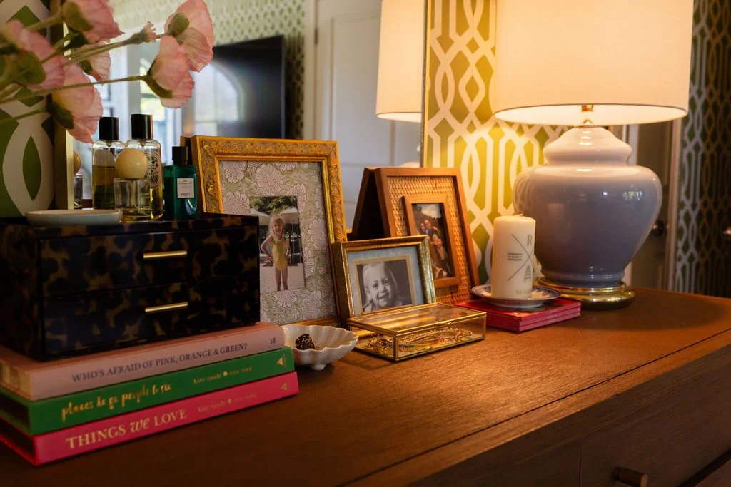 A wooden sideboard decorated with framed family photos, a white ceramic lamp, a small candles, perfume bottles, and magazines, with a large window, green and white patterned wallpaper, and pink flowers nearby.