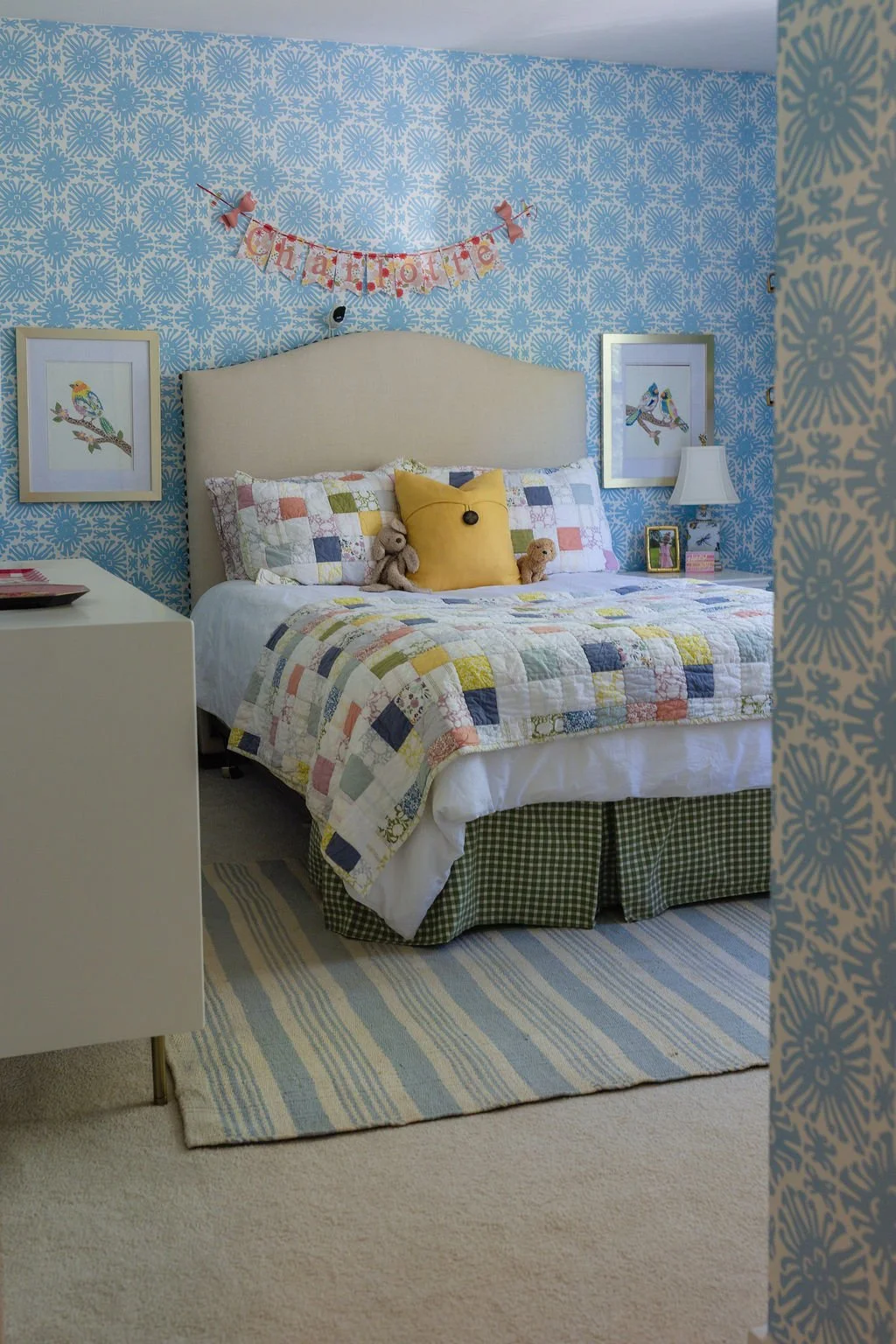 A bedroom with blue patterned wallpaper, a quilted bed with colorful patchwork bedding, and decorative pillows. Two framed bird pictures hang on the wall, and a banner with the words 'chatterbox' is above the bed. Small teddy bears sit on the bed.