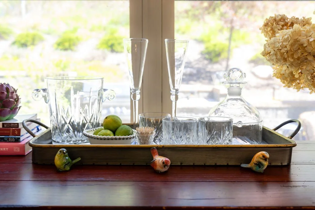 Decorative glassware on a tray, including vases, glasses, a decanter, and bowl with limes, on a wooden table near a window with a blurred outdoor view, with decorative birds on the table edges.