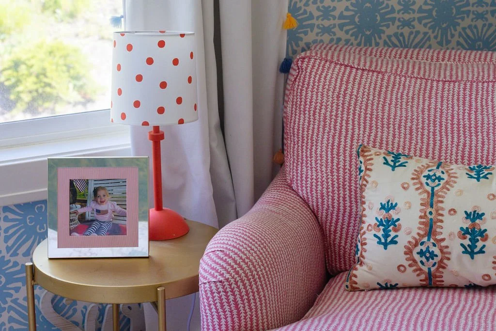 Colorful interior with a pink and white patterned armchair, pillow, a small side table with a photo frame of a girl, and a polka dot lampshade near a window.